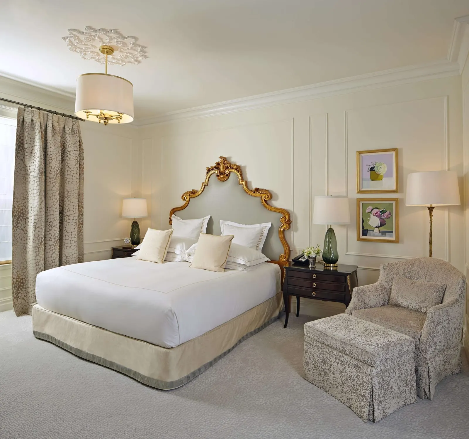 Vanderbilt Fifth Avenue Two Bedroom Suite at The Plaza: a bed with a gilded headboard on pale grey upholstery, a green glass lamp each side, floral paintings and an armchair with a footstool