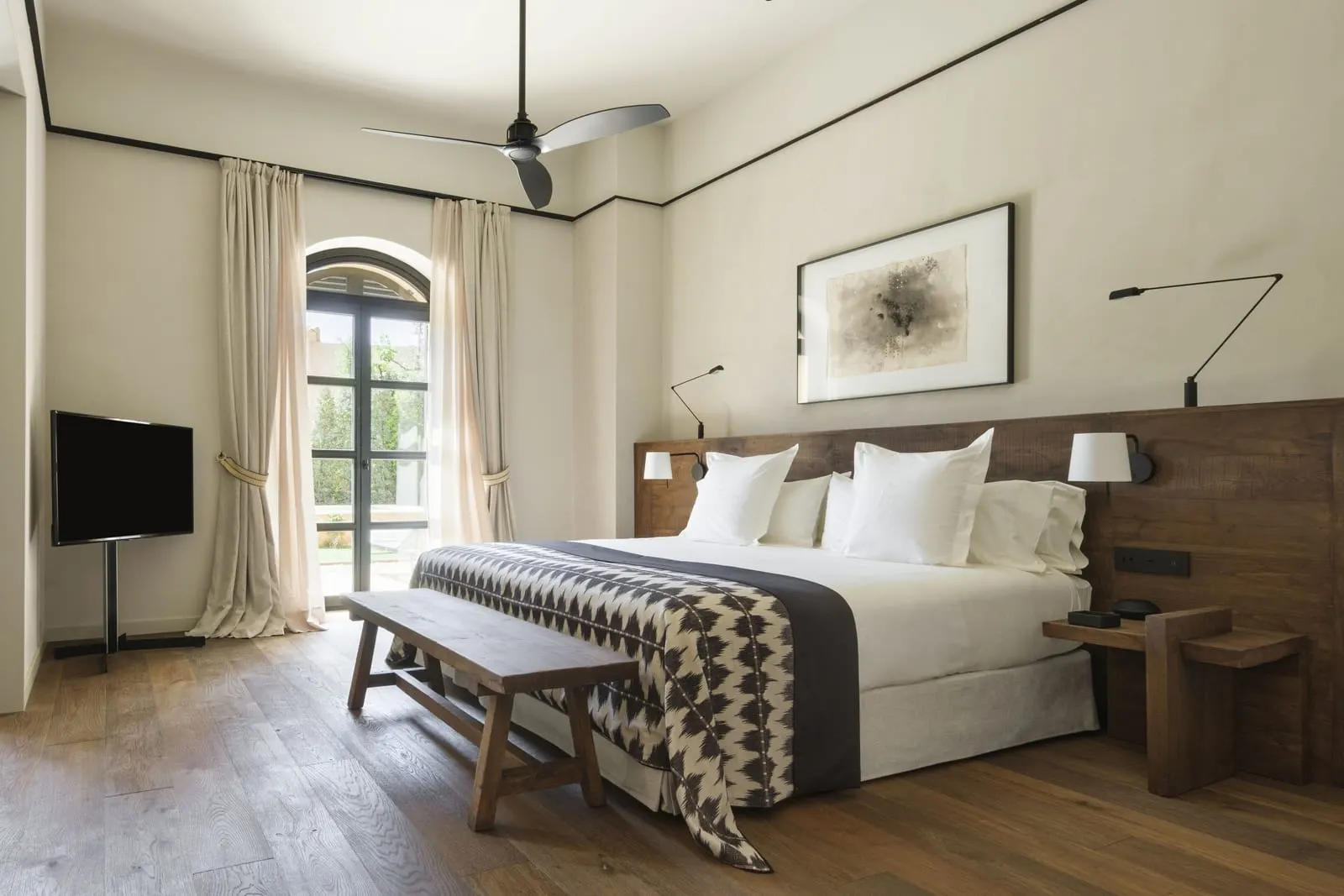 Pool Suite at Can Ferrereta: a large bed with a dark wooden headboard and a bench at its foot, a ceiling fan and an arched glass door onto a terrace and greenery