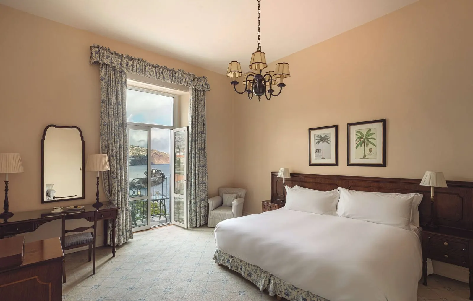 Deluxe Sea View Room at Reid's Palace, A Belmond Hotel: a large bed, a writing desk and a chandelier, with open doors to a balcony over the bay
