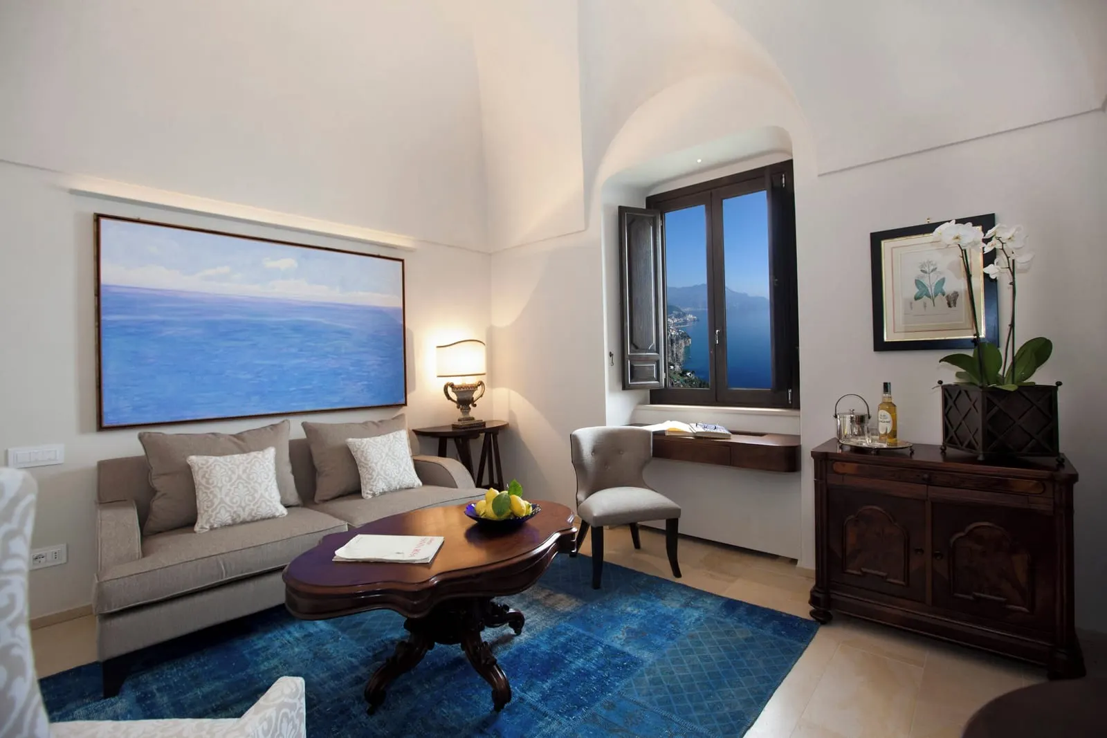 Deluxe Suite at Monastero Santa Rosa: a sitting room under a vaulted ceiling with a grey sofa, a dark wood coffee table and chest, and a blue rug.