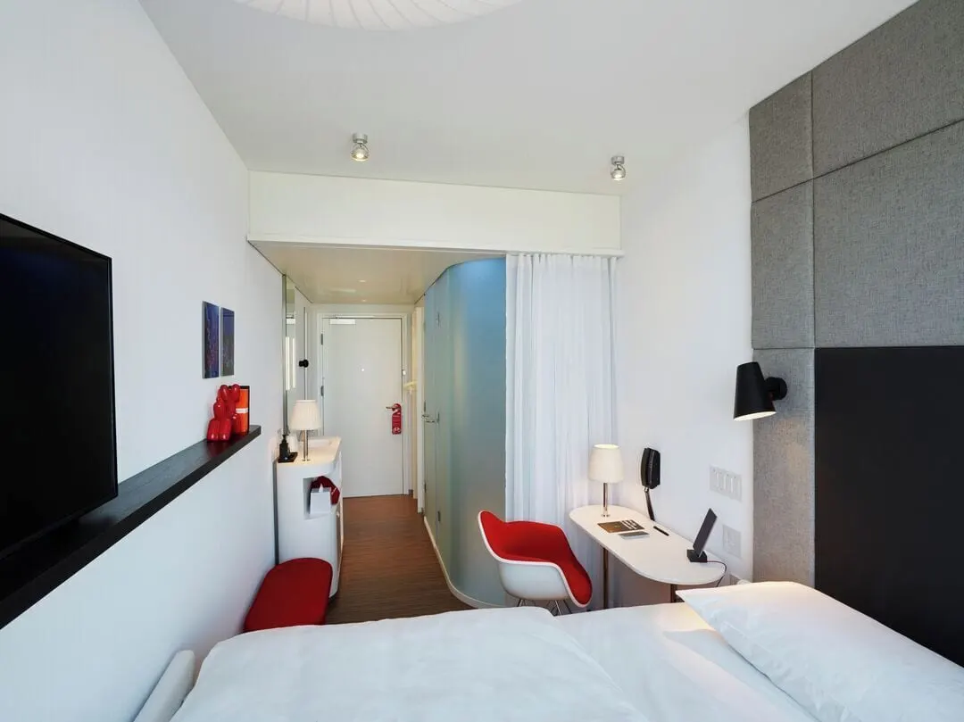 Guest room, 1 King at citizenM New York Bowery: a king bed in the foreground, with a red armchair, a small white desk, a wall-mounted TV and a frosted glass shower pod leading back to the door