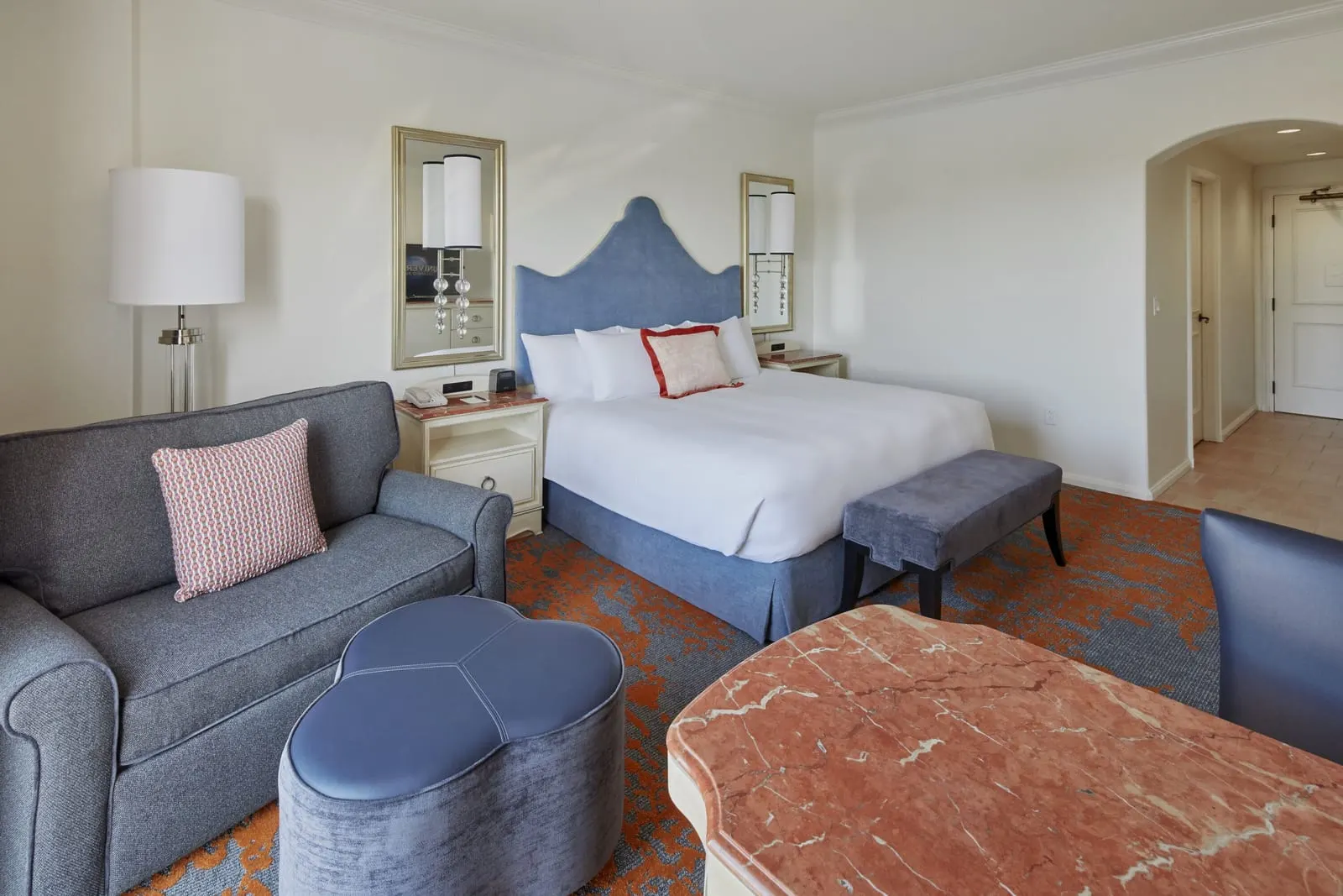 Deluxe King Room at Loews Portofino Bay Hotel: a king bed with a blue upholstered headboard, a wide armchair and ottoman, a marble-topped table and an arched hallway leading to the door