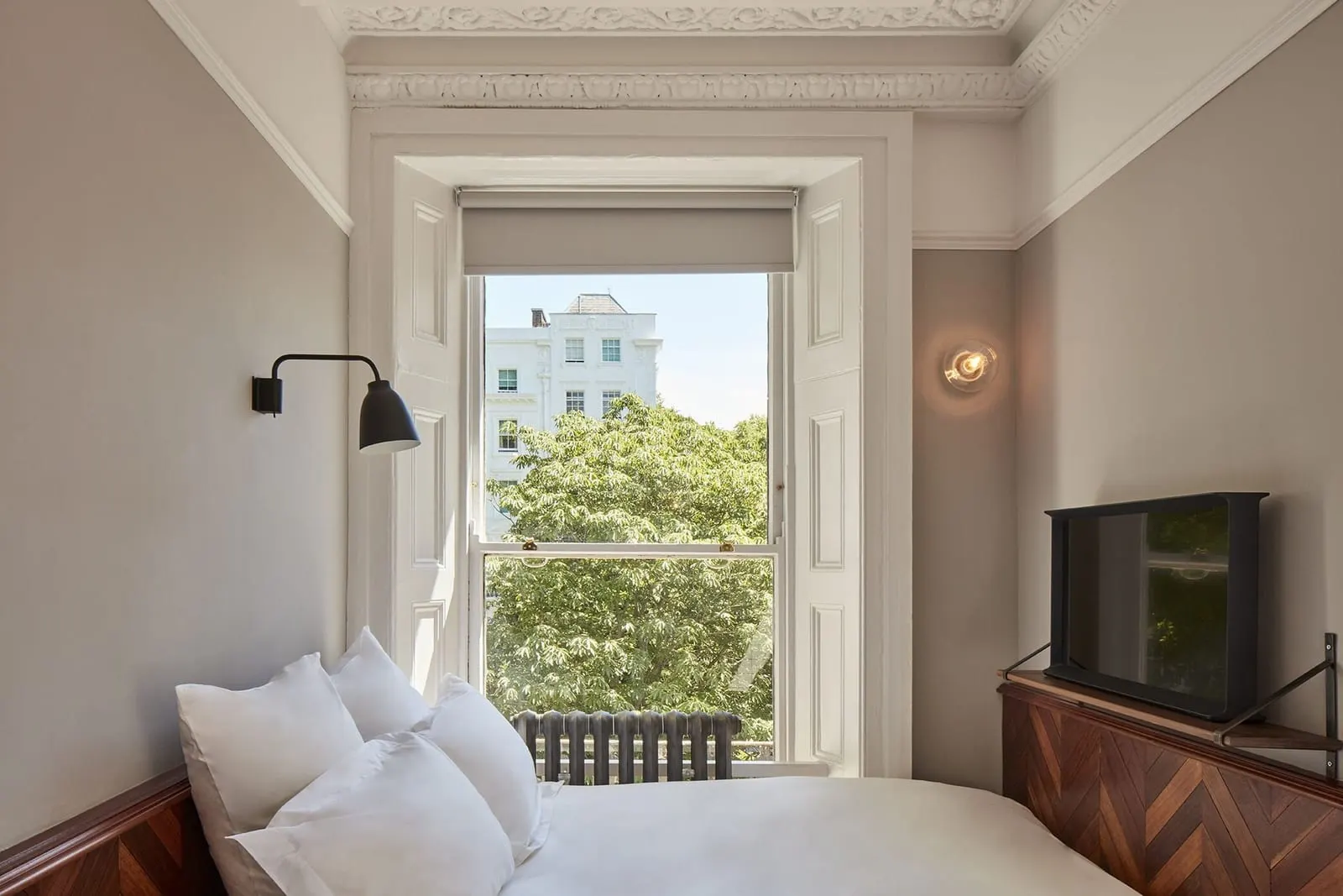 Nook at The Pilgrm: the bed, a sash window onto trees, a wall lamp and a television on a wooden unit