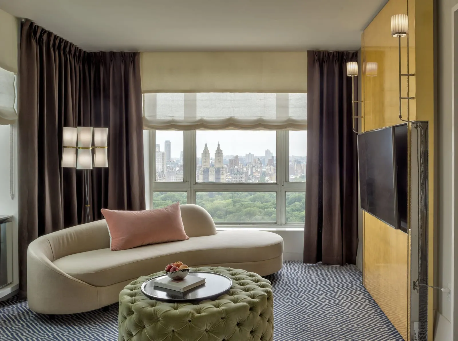 Central Park Suite at The Carlyle: a curved cream sofa and a tufted green ottoman in front of a window onto Central Park and the skyline beyond