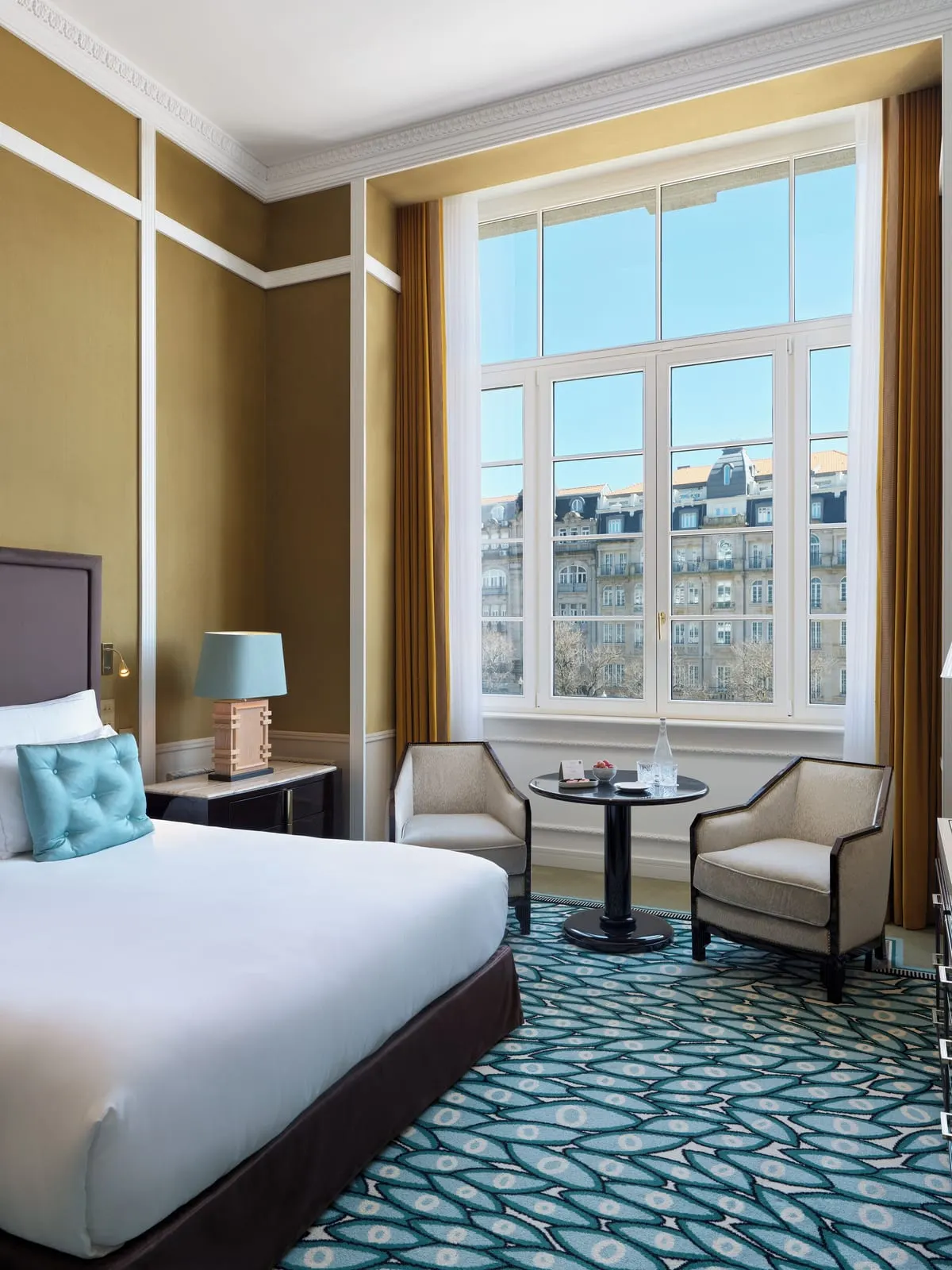 Grand Deluxe City Views room at The One Monumental Palace: the bed, two armchairs and a tall window onto the facades opposite