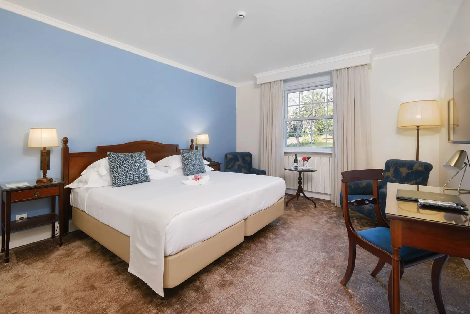 Double Garden Wing at Casa Velha do Palheiro, Relais & Châteaux: a king bed made from two twins against a blue wall, a desk and armchairs by a sash window over the garden