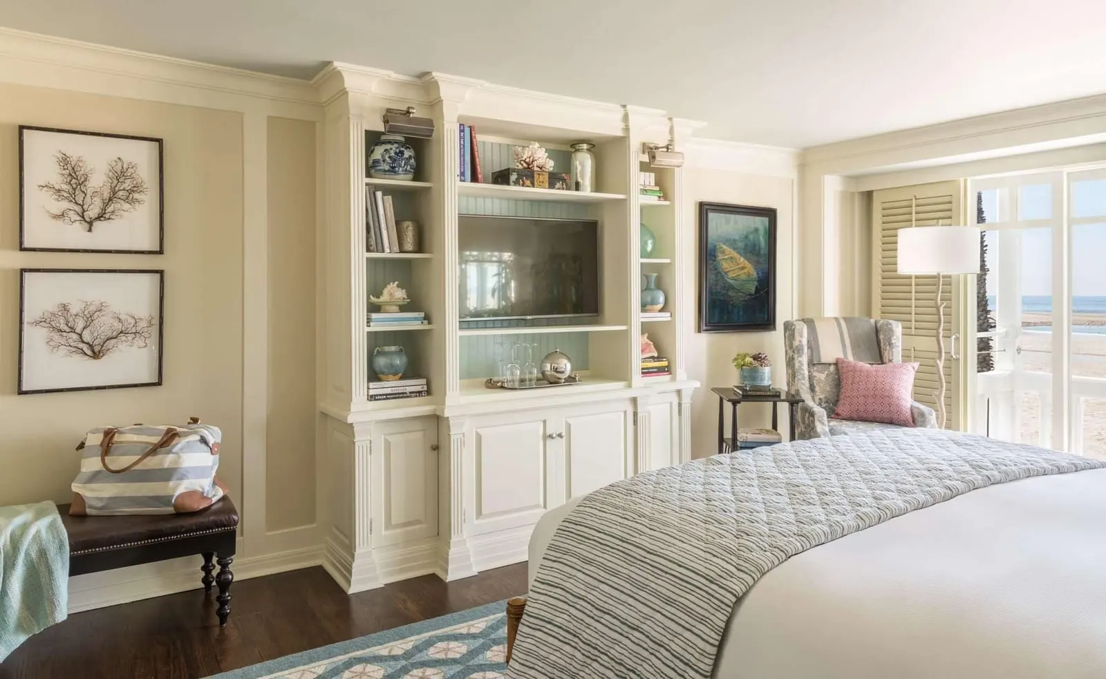Ocean View Room at Shutters on the Beach: a bed beside a cream built-in unit with a television and shelves, a wing armchair, and glass doors to a balcony with the beach and the sea beyond