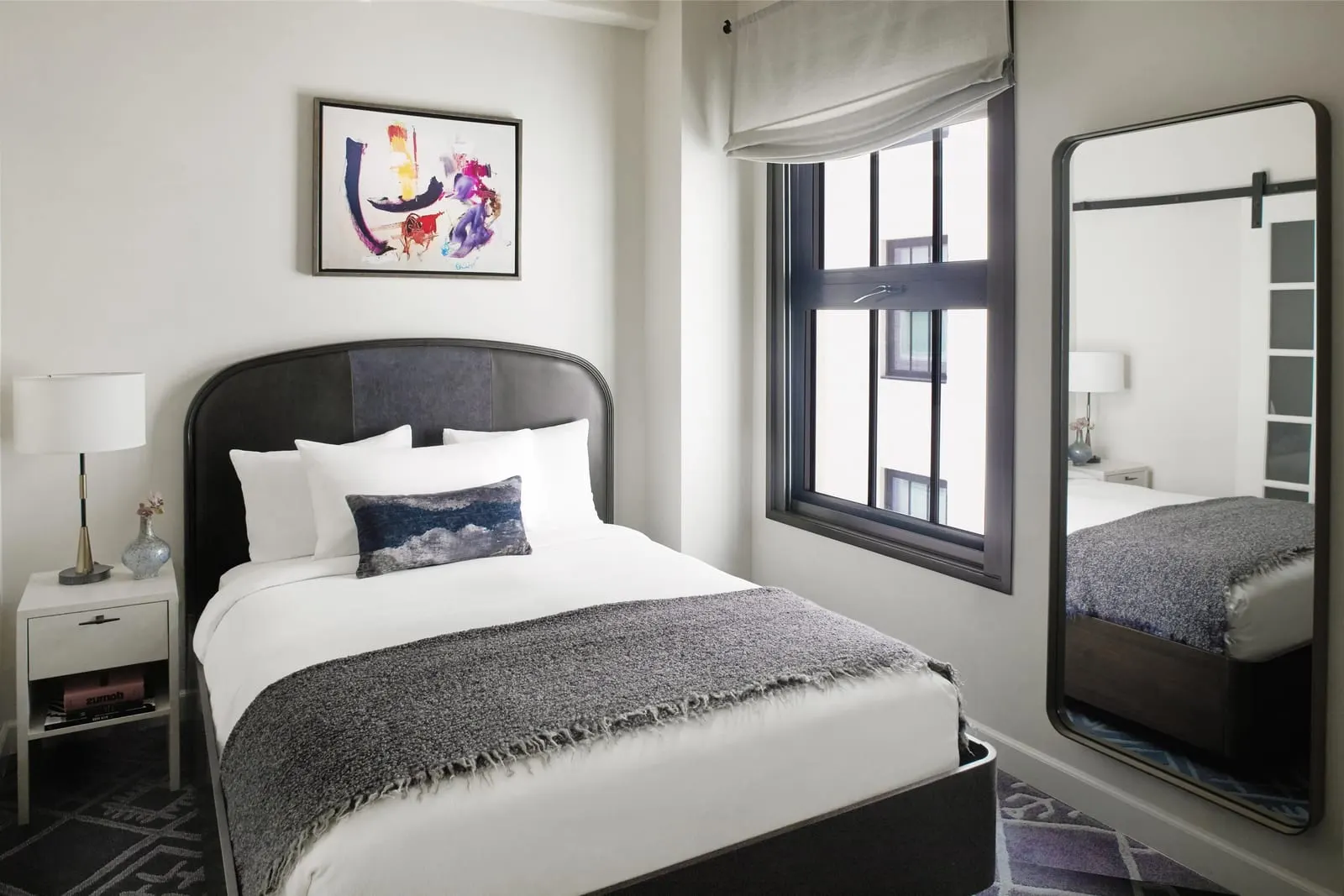 1 Queen Bed im Hotel Figueroa: ein Queensize-Bett mit einem dunklen geschwungenen Lederkopfteil, ein weißer Nachttisch mit Lampe, ein abstraktes Gemälde und ein hoher Spiegel neben einem Schiebefenster