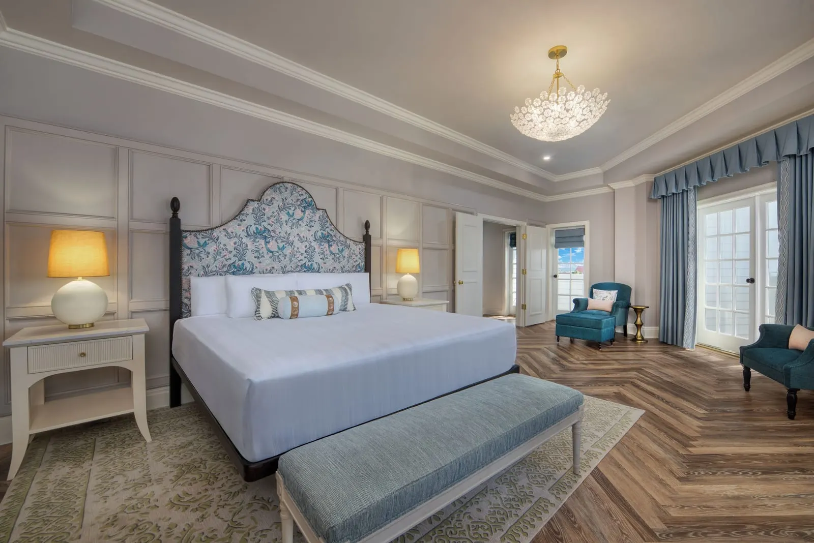 Victorian Suite - Club Level at Disney's Grand Floridian Resort & Spa: a king bed with a blue and white patterned headboard and a padded bench, with two armchairs by curtained French doors and a herringbone wood floor
