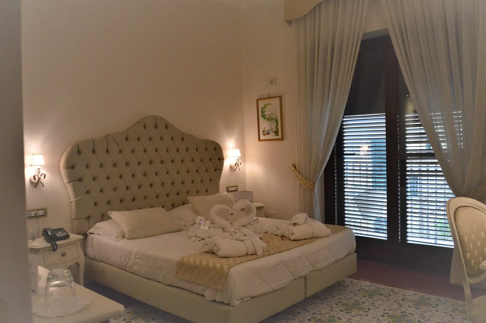 De Luxe Room at Hotel Santa Lucia: a large bed with a tall buttoned headboard beside curtained doors with shutters