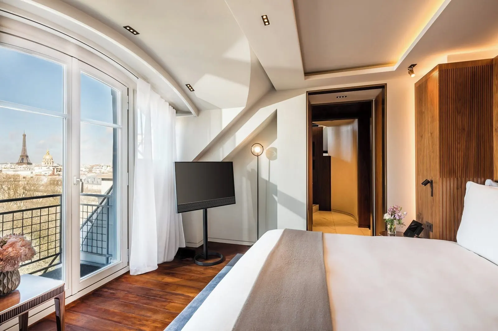 Amour Suite at Mandarin Oriental Lutetia, Paris: a double bed facing a curved French window onto a balcony, with the Eiffel Tower and the gilded dome of Les Invalides beyond, and a sloping ceiling over a television