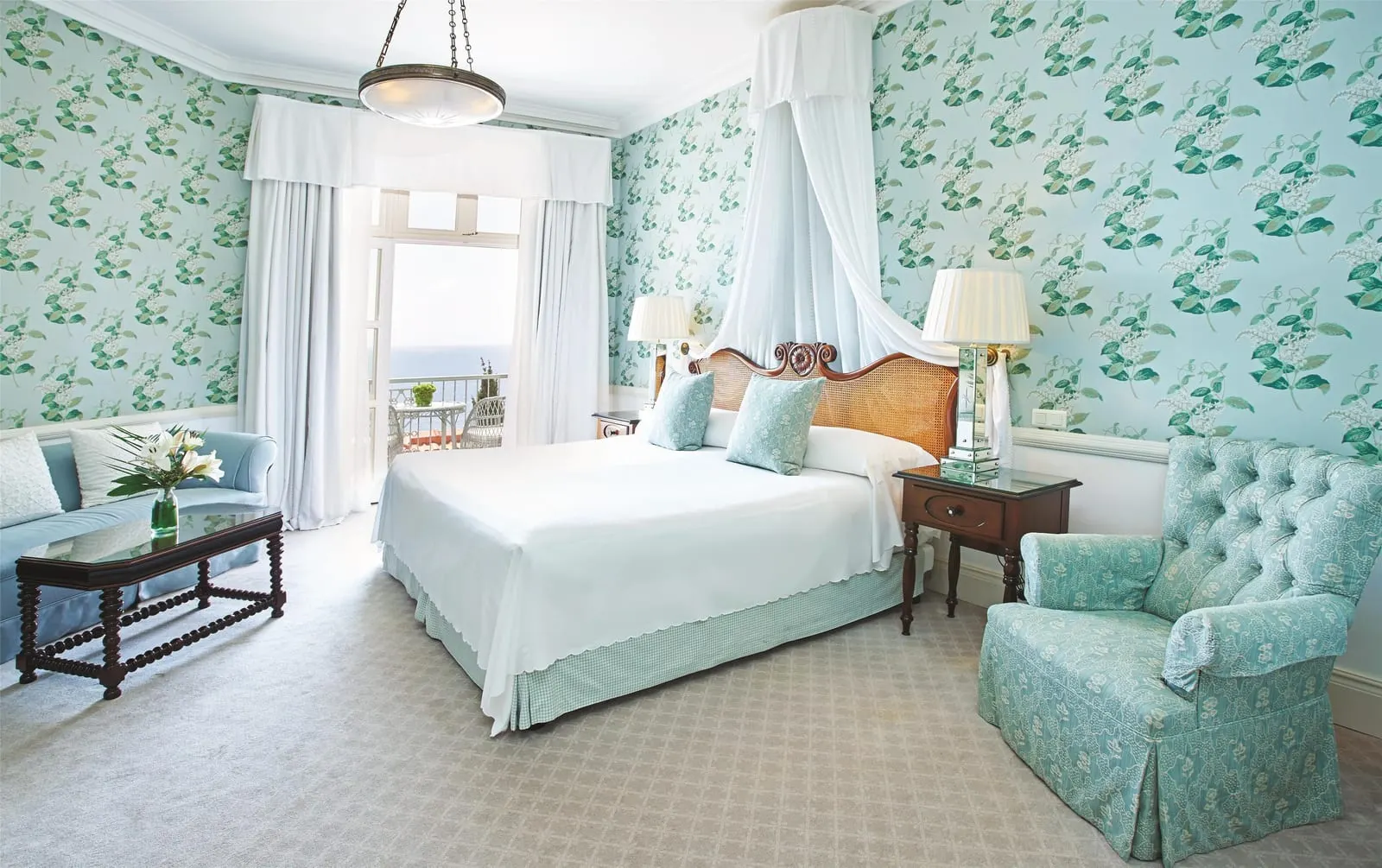 Deluxe Junior Suite at Reid's Palace, A Belmond Hotel: a bed under a white canopy, leaf-patterned wallpaper and a sofa and armchair by open balcony doors
