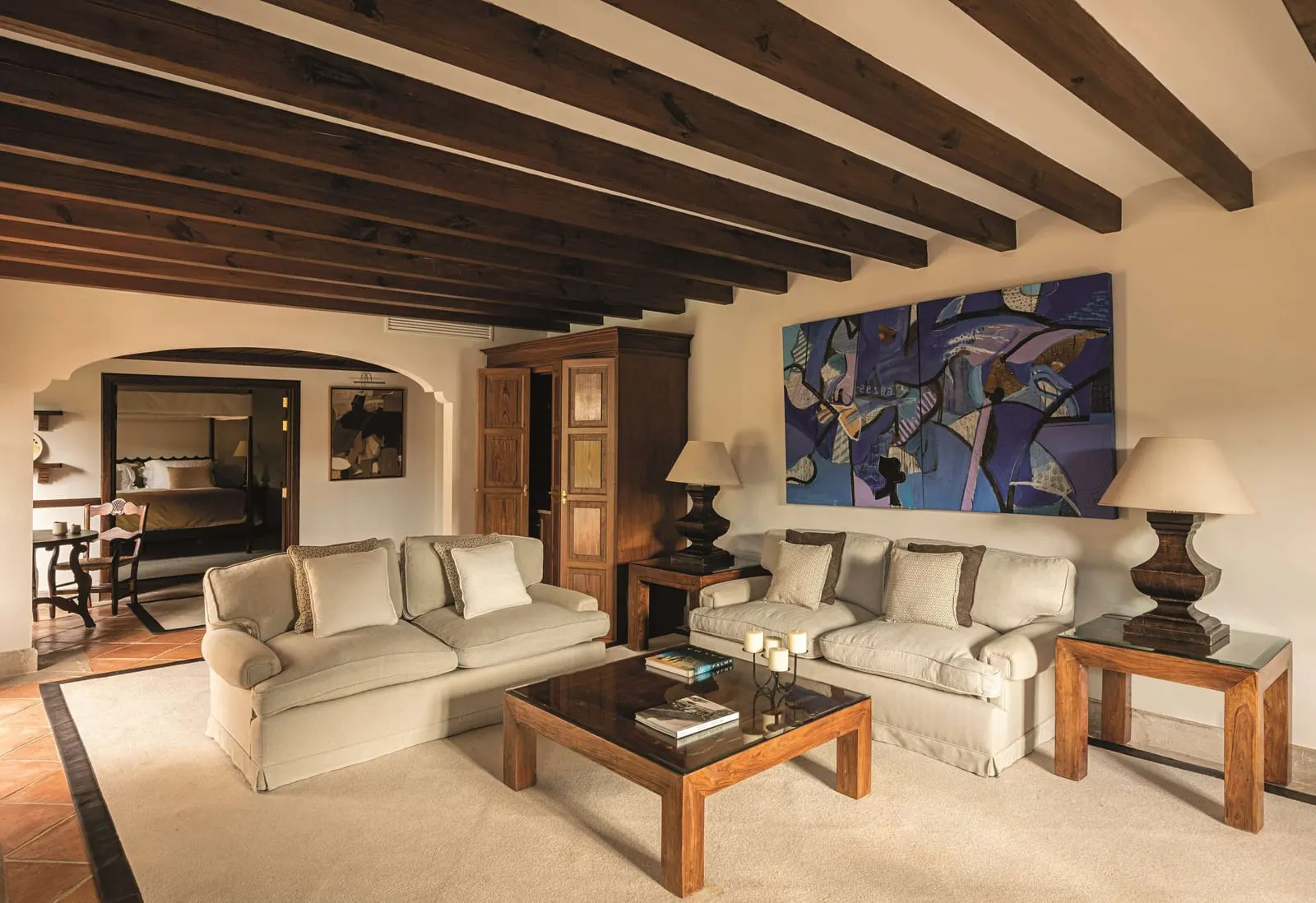 Sheridan Suite at Belmond La Residencia: a sitting room with two cream sofas around a glass-topped coffee table under dark beams, a large blue abstract artwork and an arch to a four-poster bedroom