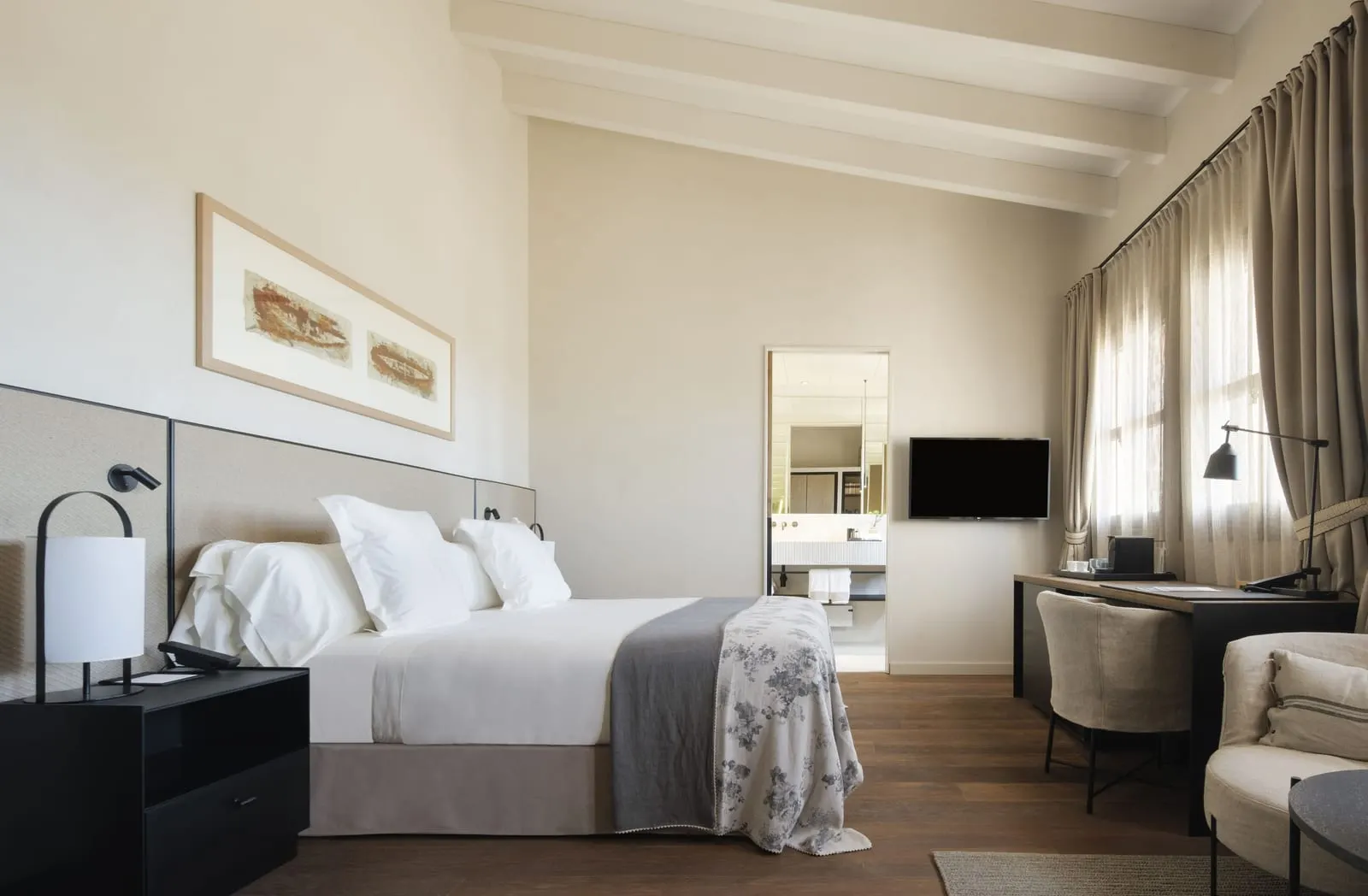 Deluxe Room at Can Ferrereta: a bed under pale beams on a sloping ceiling, a wooden floor, a desk by the curtained windows and the bathroom visible through an open door
