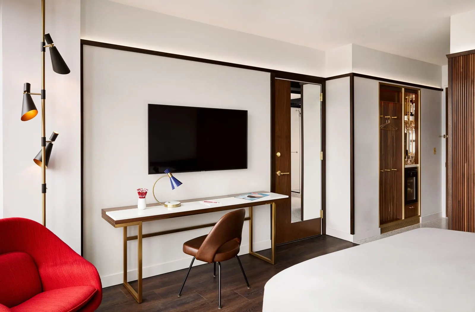 Deluxe King at TWA Hotel: a long white-topped desk under a wall-mounted television, a brown leather chair and a red armchair, with the corner of the bed in the foreground