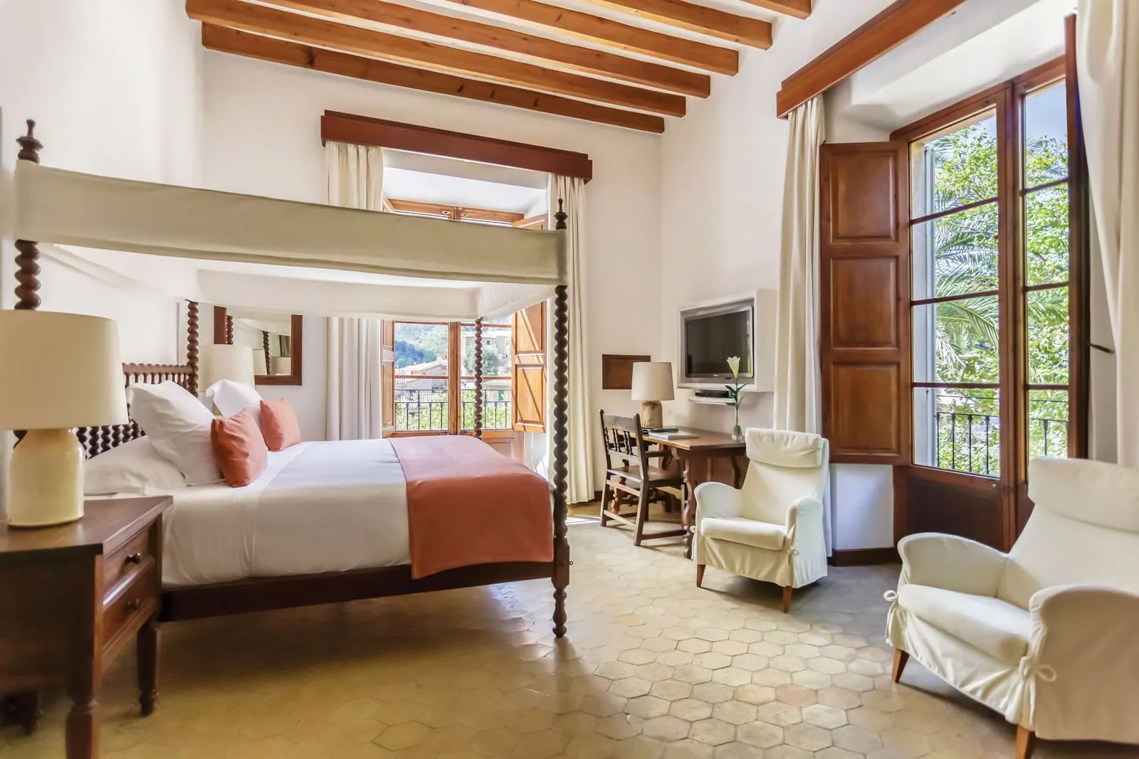 Deluxe Double Room at Belmond La Residencia: a four-poster bed with a white canopy under wooden beams on a tiled floor, with a desk, two white armchairs and tall shuttered windows