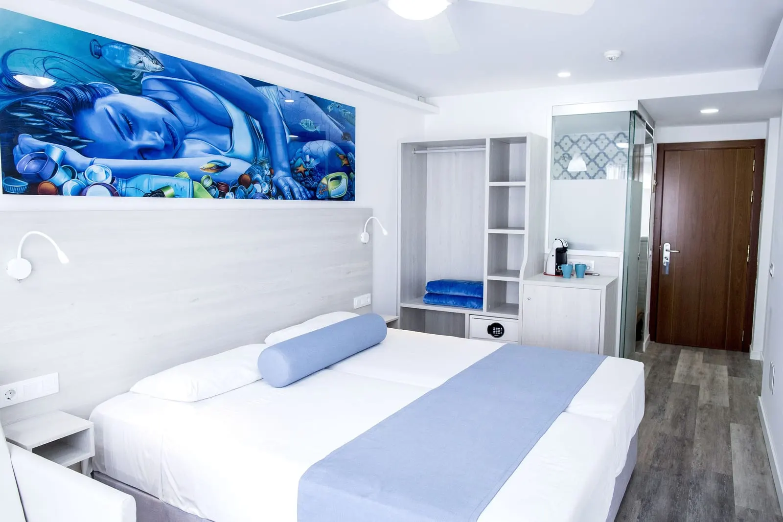 Surfing Room（Corralejo Surfing Colors Hotel & Apartments）：大きな青い壁画の下に2台のシングルベッドを寄せて配置し、オープンタイプの棚式ワードローブとシーリングファンがある