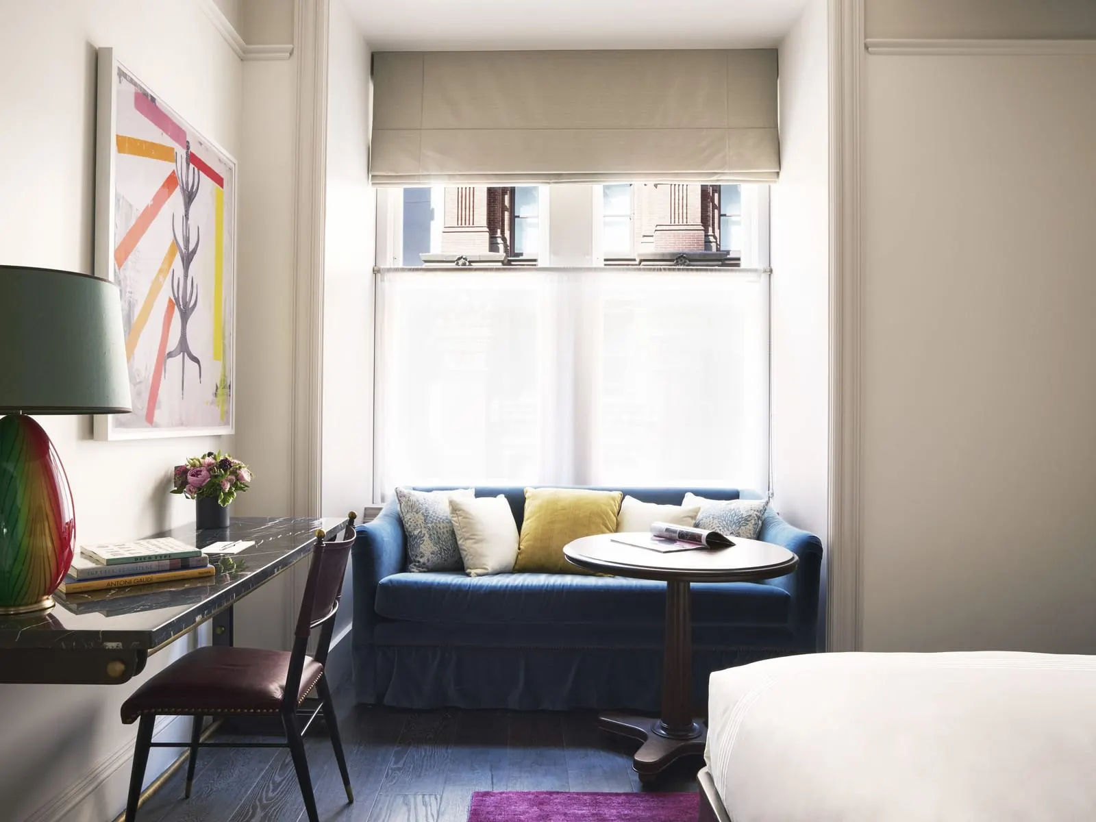 Premium King at The Beekman: a blue velvet sofa and a round wooden table in a window nook, with a dark marble desk, a coloured glass lamp and abstract art on the left