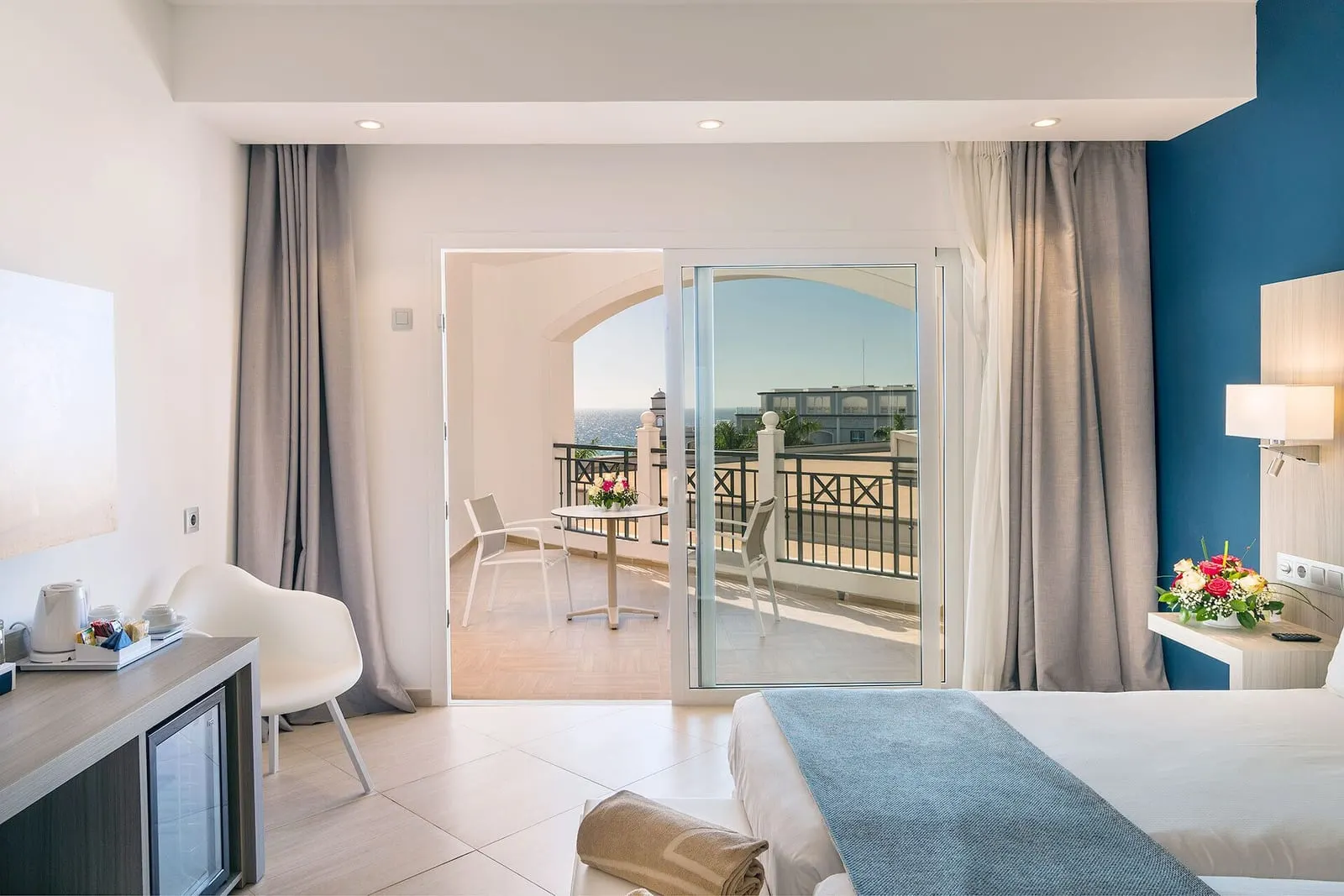 Double Lateral Sea View at Royal Palm Resort & Spa: the foot of the bed and a minibar unit, with glass doors open to a balcony with a table and chairs