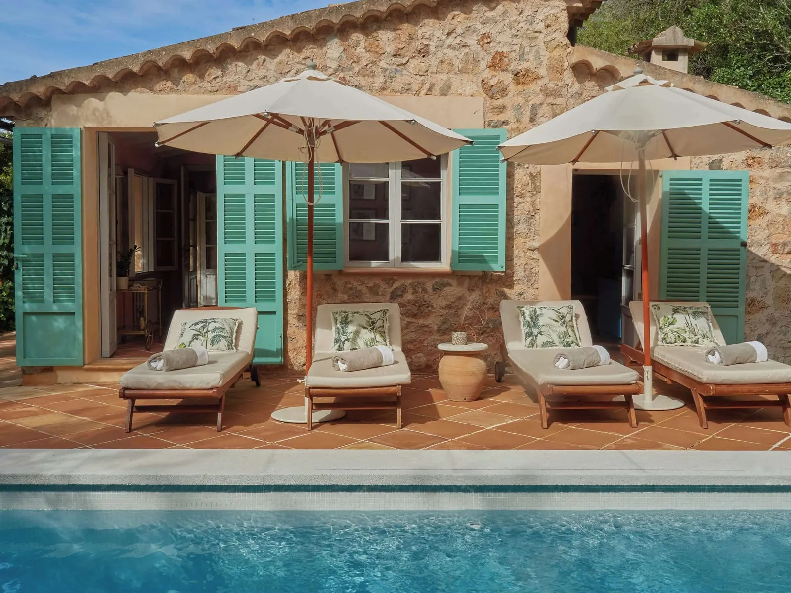 Matthew Williamson Suite at Belmond La Residencia: four sunloungers and two parasols on a tiled terrace beside a pool, in front of a stone building with turquoise shutters