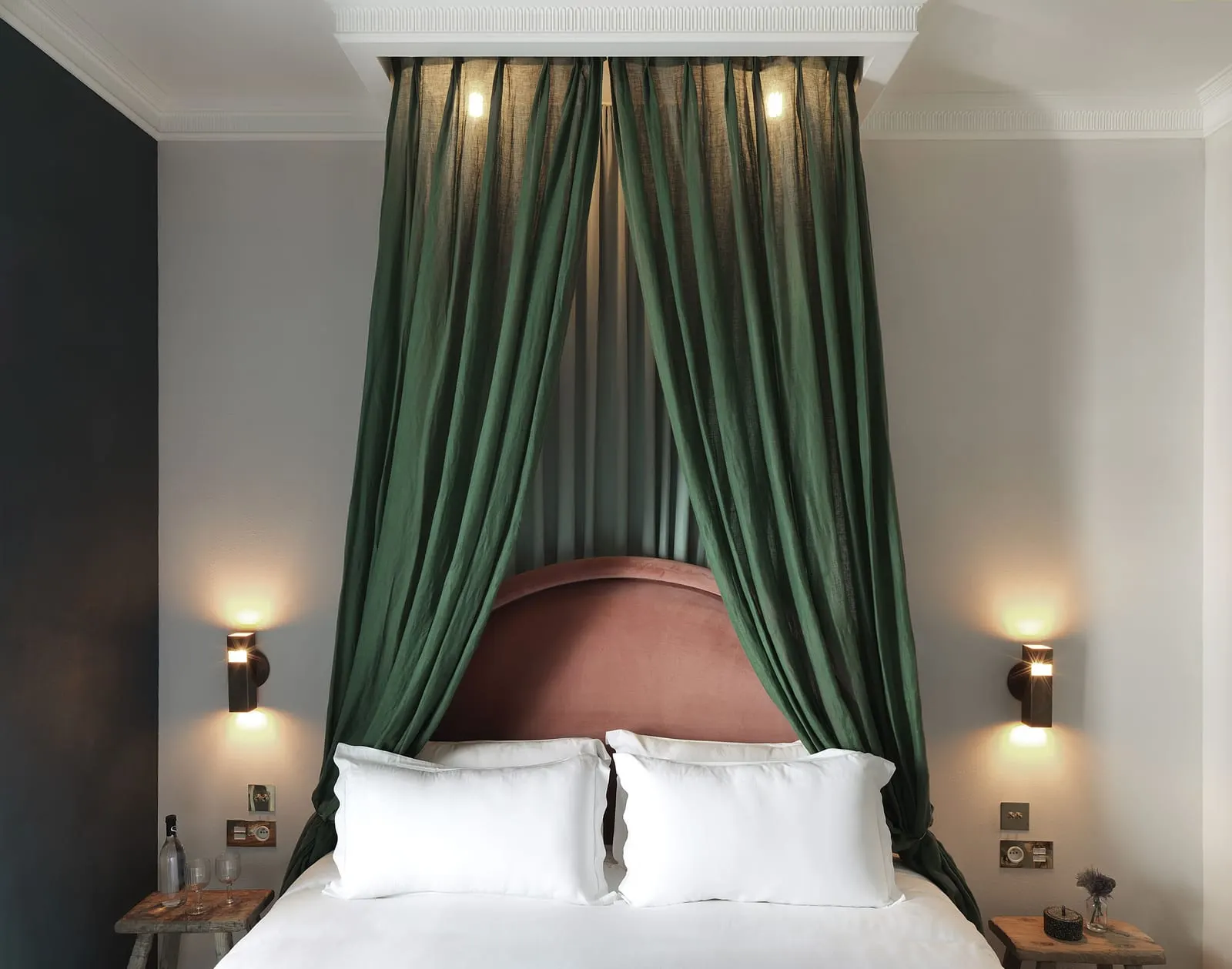 Petit Boulevard Balcony at Grands Boulevards Experimental: a bed under a dark green canopy hung from a moulded ceiling box, with a pink velvet headboard, wall lights on each side and wooden stools as bedside tables