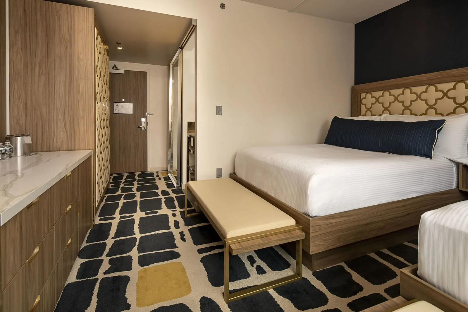 Blanca Double at SAHARA Las Vegas: a bed with a quatrefoil headboard and a bench at its foot, the end of a second bed, a long wooden dresser with a marble-effect top and the hallway to the door