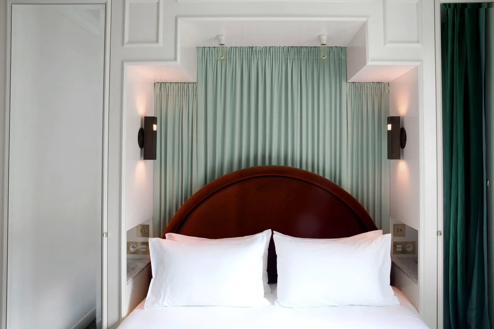 Petit Boulevard Single at Grands Boulevards Experimental: a bed with a rust velvet headboard set into a white panelled alcove hung with pale green curtains, with a wall light and a marble ledge on each side