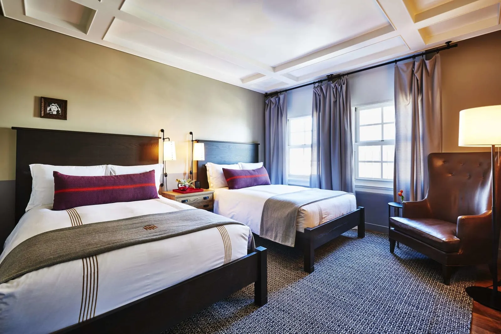 Tower Room im The Hollywood Roosevelt: zwei Doppelbetten mit dunklen Kopfteilen und grauen Tagesdecken, ein Ledersessel und hohe Schiebefenster mit graublauen Vorhängen