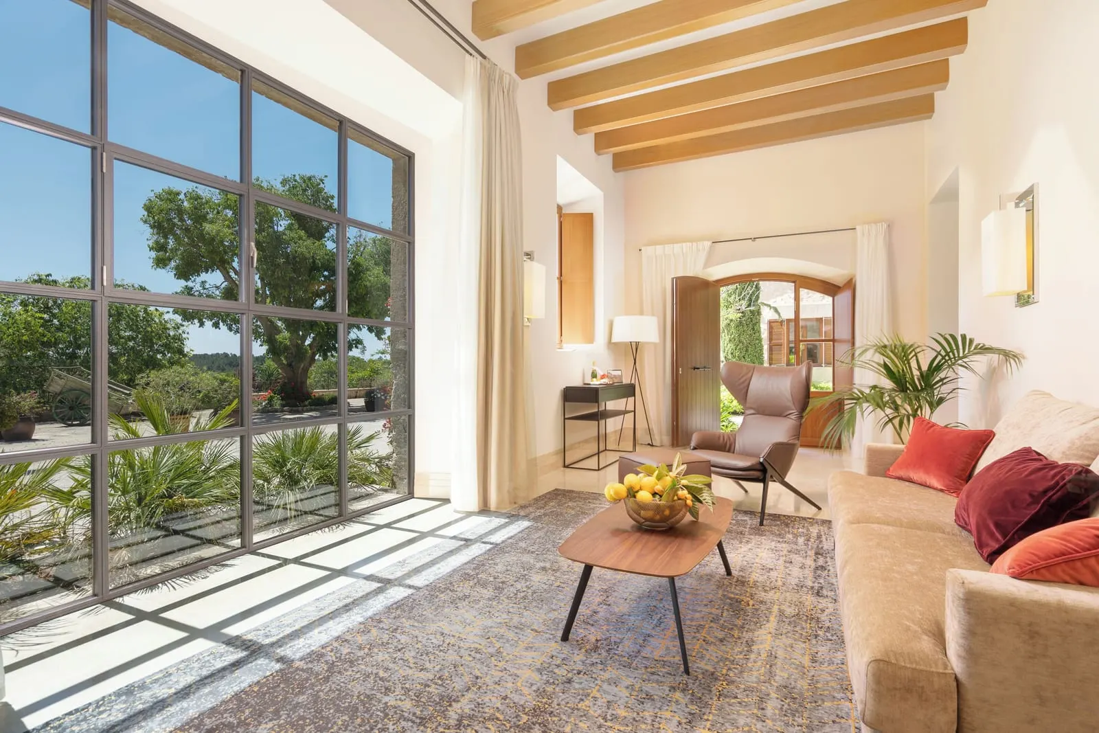 Castell Suite im Castell Son Claret: ein Wohnzimmer mit einem Sofa, einem Ledersessel und einem niedrigen Tisch neben einem hohen Fenster mit Stahlrahmen, unter einer Decke mit Holzbalken