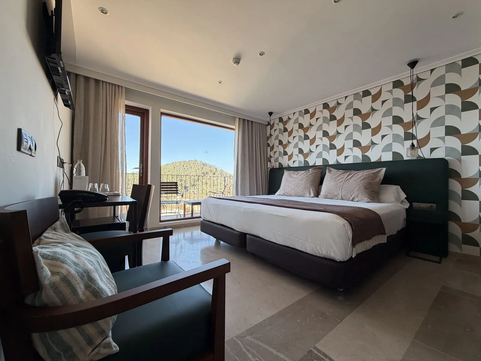 Dreaming Sea at Maristel Hotel & Spa: a large bed against a patterned wall, an armchair and a desk, with a tall window onto a balcony and a wooded hillside