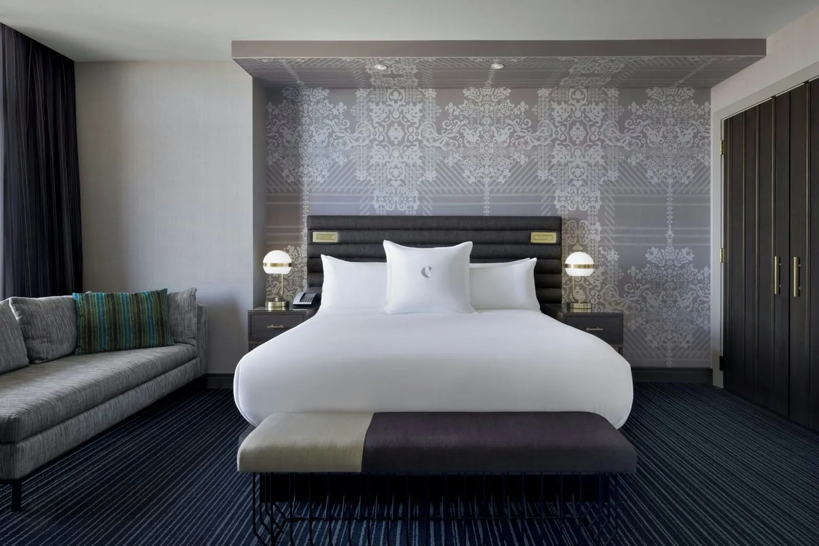 Terrace Suite Fountain View at The Cosmopolitan of Las Vegas: the bedroom, with a king bed against a patterned wall, a grey sofa and a wall of dark wardrobe doors