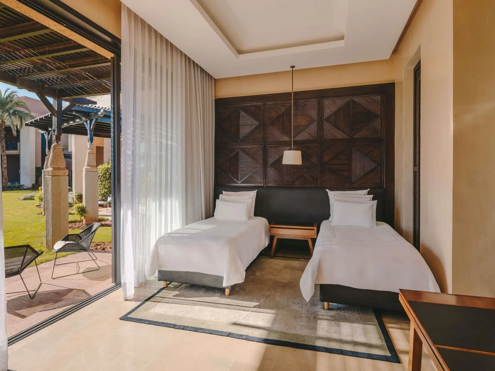 Family Suite im Fairmont Royal Palm Marrakech: ein zweites Schlafzimmer mit Einzelbetten und einer Wand mit geschnitztem Holzkopfteil, das sich auf eine Terrasse und einen Rasen öffnet
