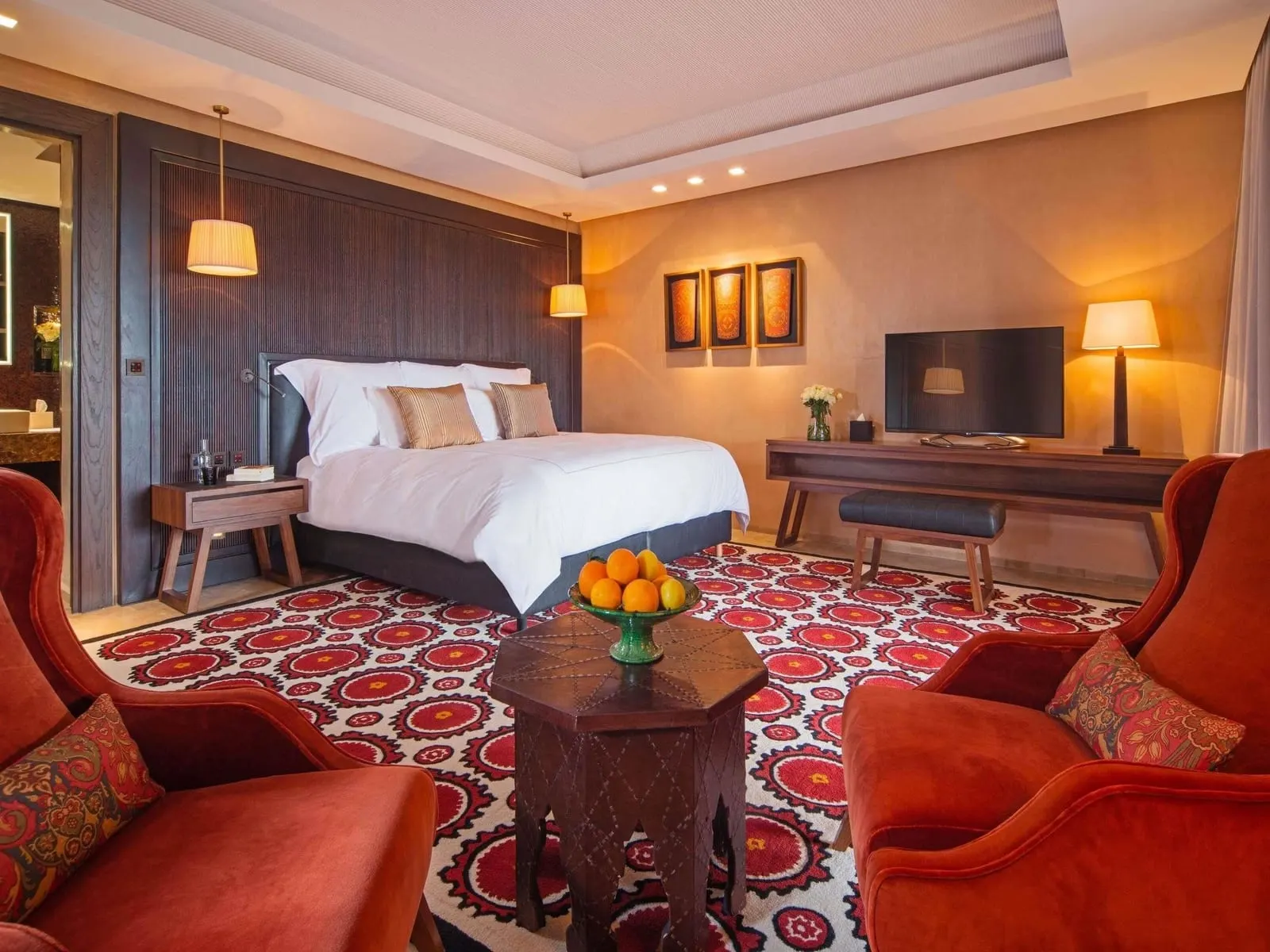 Deluxe Suite Garden View im Fairmont Royal Palm Marrakech: ein Kingsize-Bett, ein Schreibtisch und ein roter gemusterter Teppich unter warmen Lampen