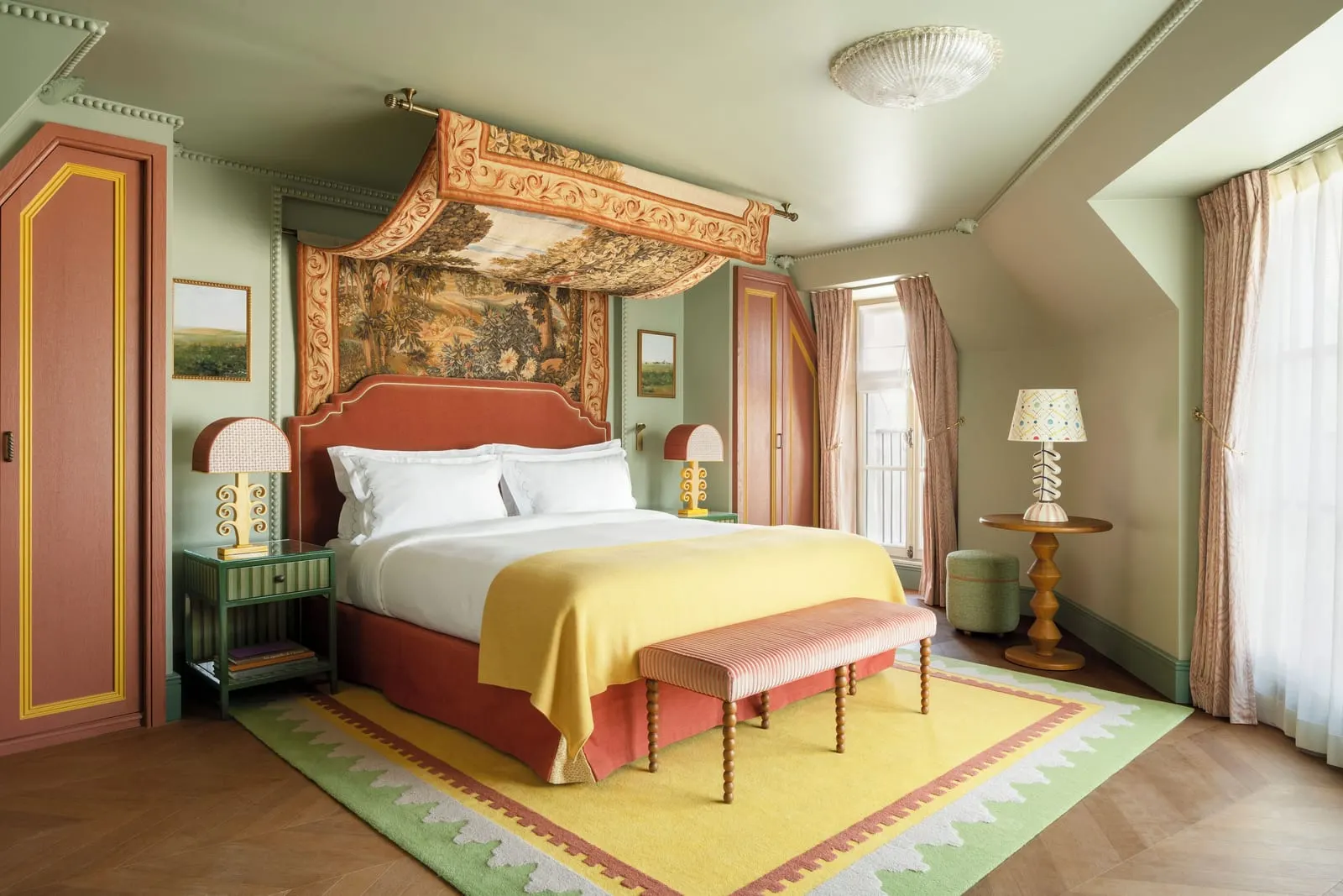 Parisian Junior Suite at Le Grand Mazarin: a bed under a tapestry canopy with a striped bench at its foot, beneath a sloping ceiling with painted cupboards and tall windows