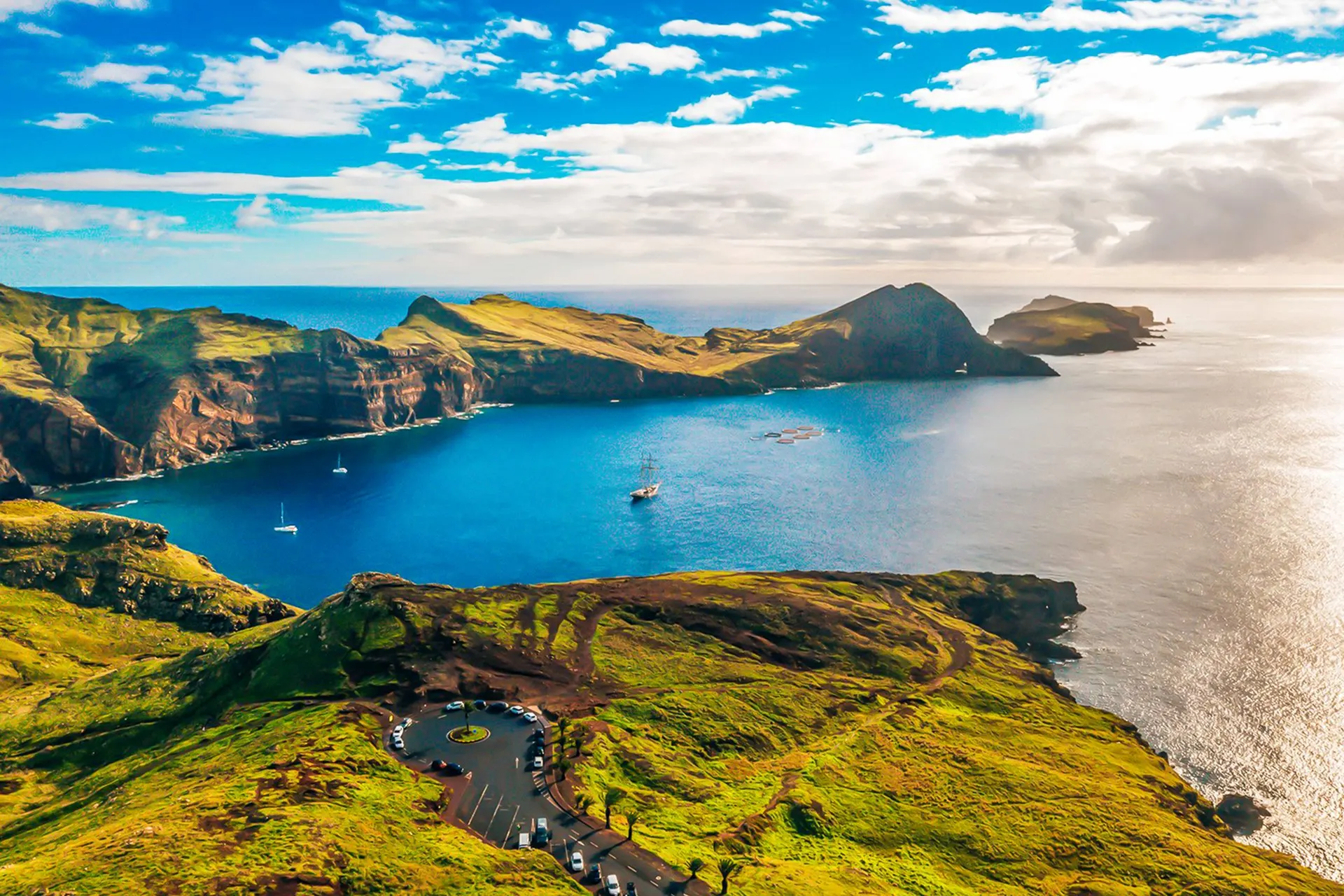 The Ponta de São Lourenço headland stretching east into the Atlantic, green ridges and red cliffs above a deep blue bay, with a car park at the end of the road in the foreground
