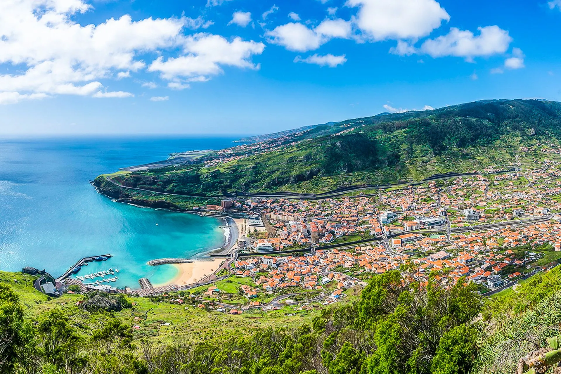 Machico bay and town from the hillside above, with the sandy beach, the harbour and the airport runway along the coast beyond