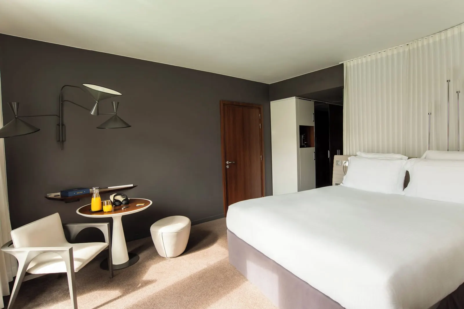 Deluxe Room at Hôtel Molitor Paris: a king-size bed, a white armchair and a small round table against a dark grey wall, with the entrance door and wardrobe behind