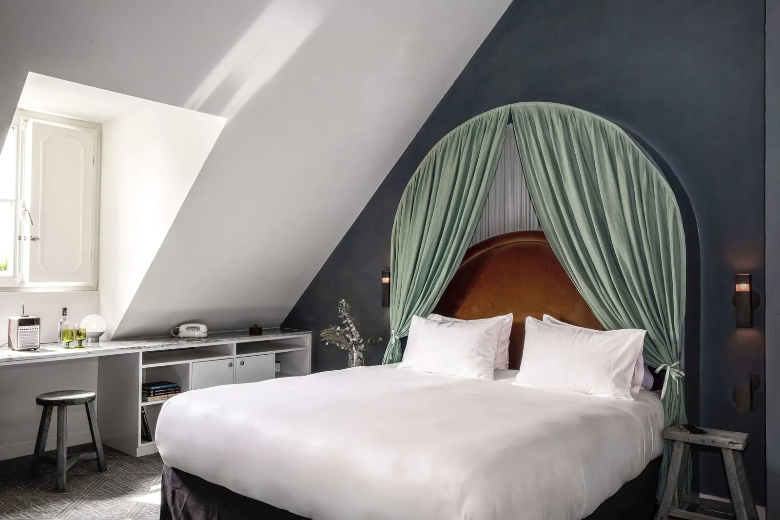 Parisian Eaves at Grands Boulevards Experimental: a bed under an arched sage green canopy with a rust velvet headboard, set into a dark blue wall under the slope of the roof, beside a dormer window and a built-in marble-topped desk with a wooden stool
