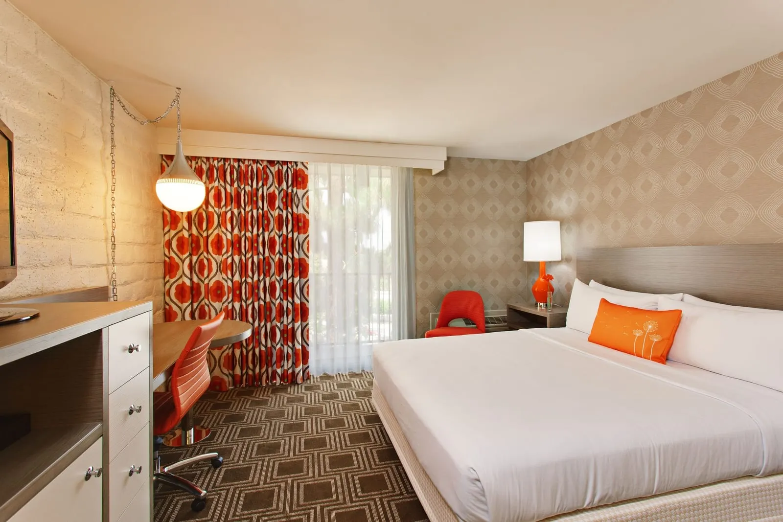 Deluxe King Pool View im The Garland: ein Kingsize-Bett mit einem orangefarbenen Kissen, ein Schreibtisch mit einem orangefarbenen Stuhl und ein Fernseher, neben einem Fenster mit orange gemusterten Vorhängen