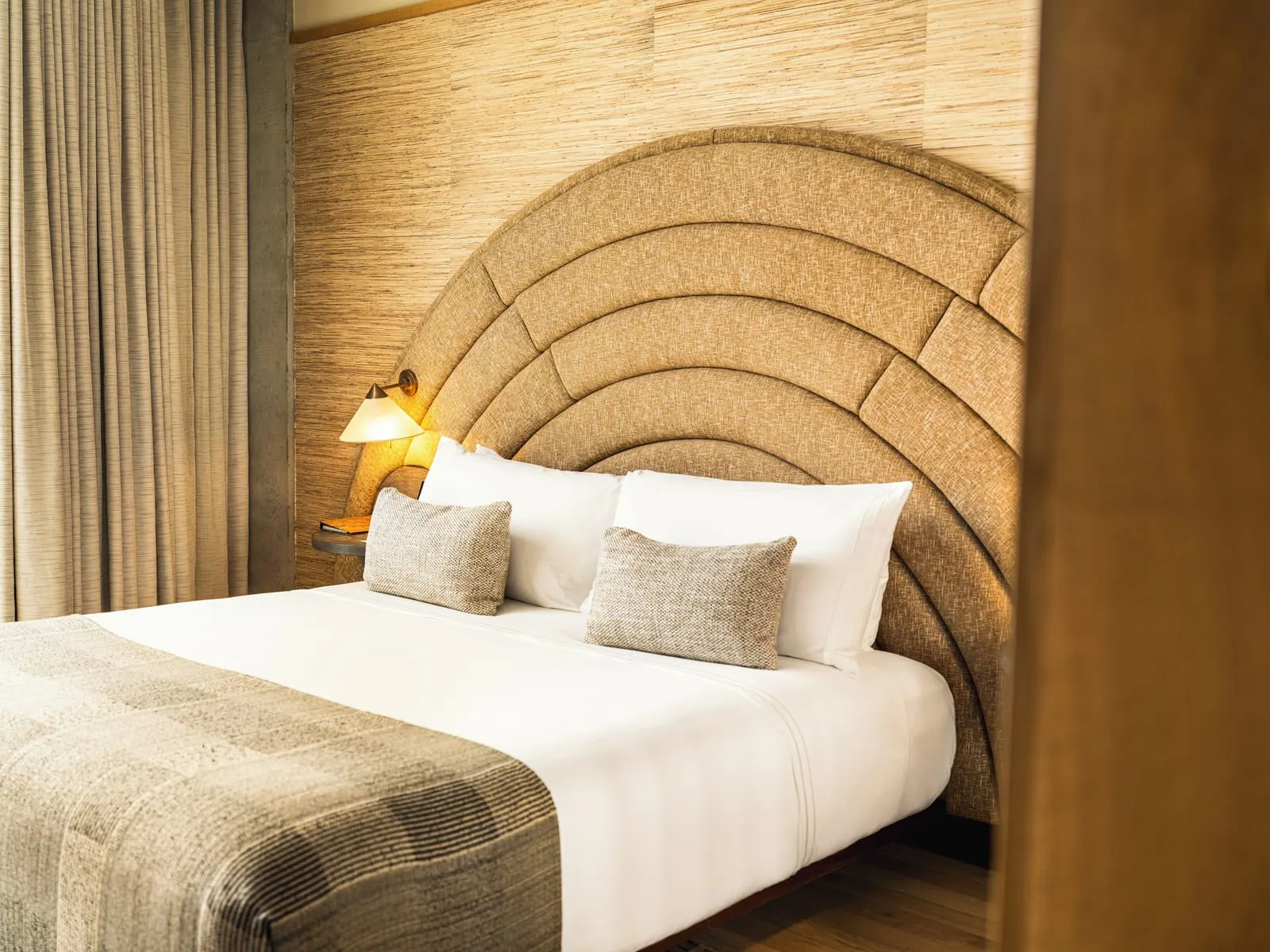 Deluxe King im Santa Monica Proper Hotel: ein Kingsize-Bett vor einem bogenförmigen Kopfteil und einer geflochtenen Wandverkleidung, mit bodenlangen Vorhängen auf einer Seite