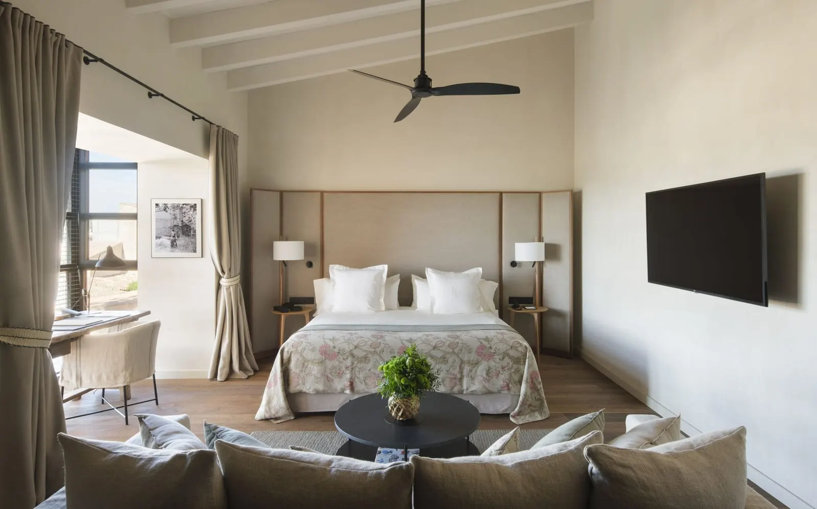 Can Ferrereta Suite at Can Ferrereta: a bed with a padded headboard under pale beams and a ceiling fan, with a sofa in the foreground and a desk by the window