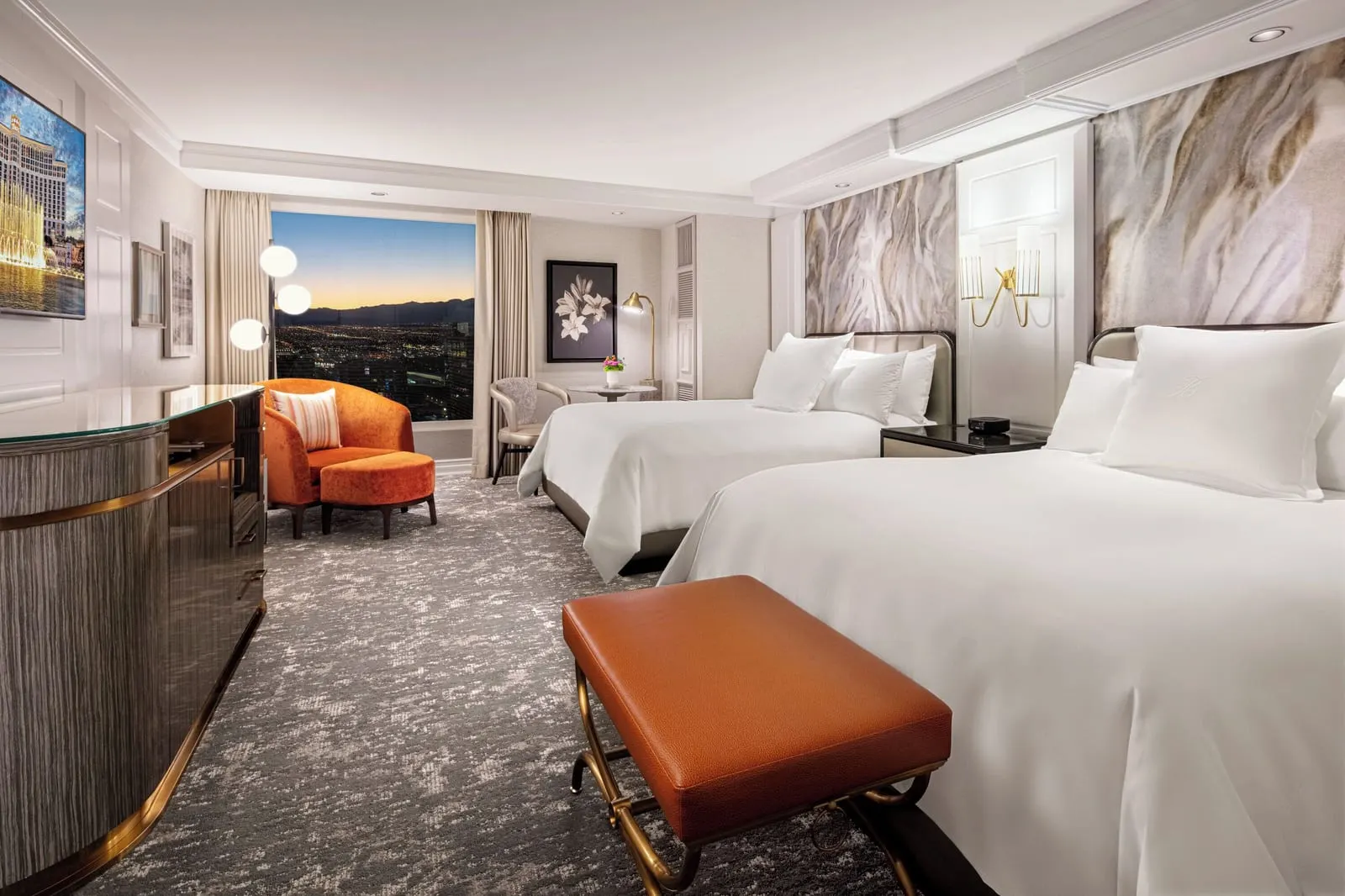 Spa Premier Two Queen at Bellagio: two queen beds with a leather bench at the foot of one, an orange armchair and footstool, and a small round table by the window