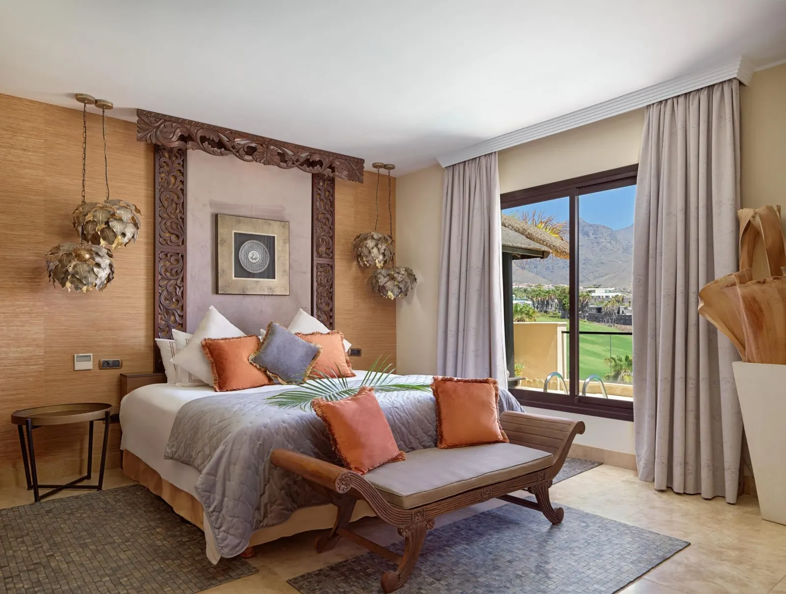 Royal Villa at Royal Garden Villas: a bedroom with orange cushions, a carved bench and a window onto palms and hills