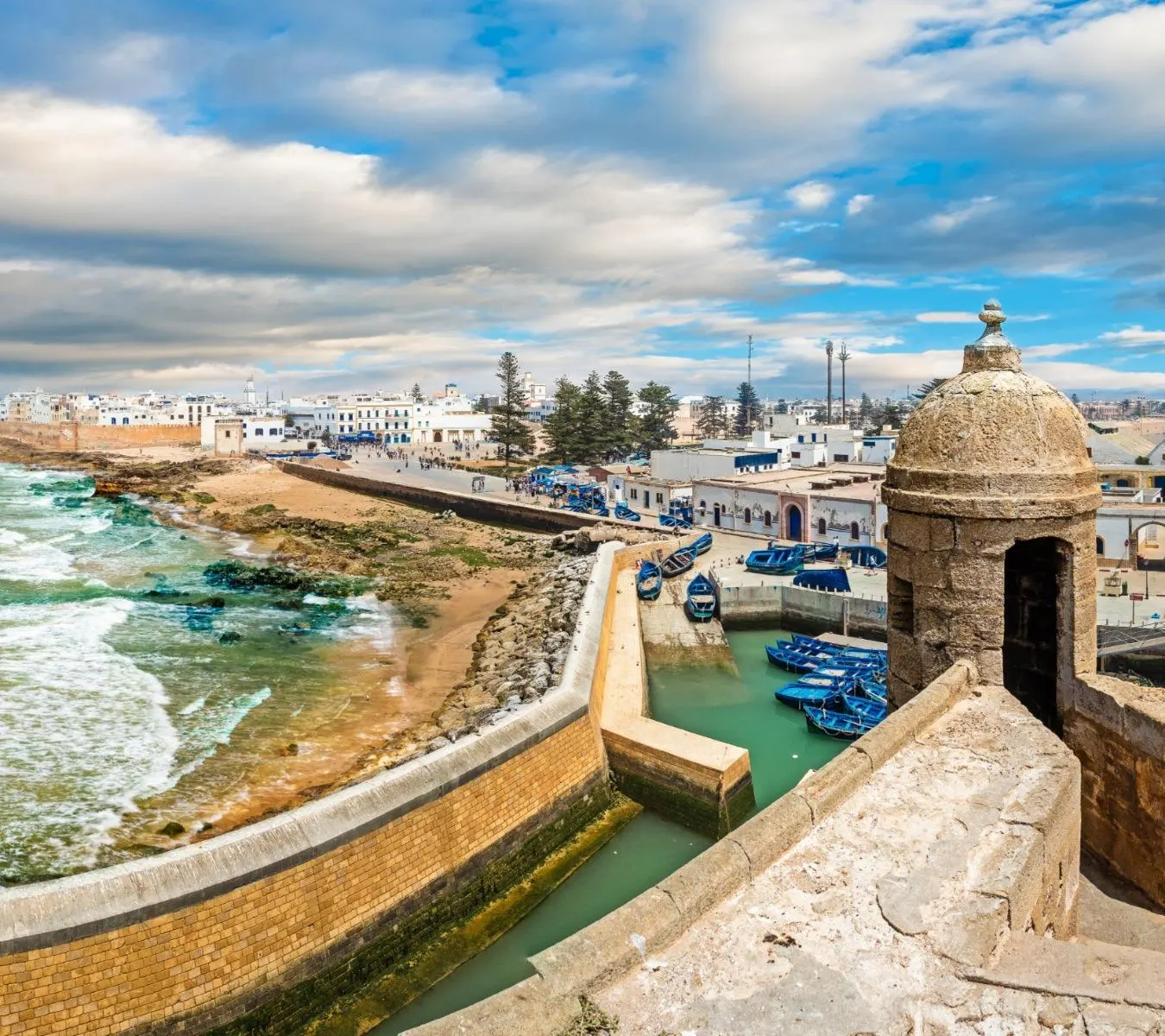 View from a stone sentry box on Essaouira's sea wall over the small boat harbour, the beach and waves on the left and the white medina beyond