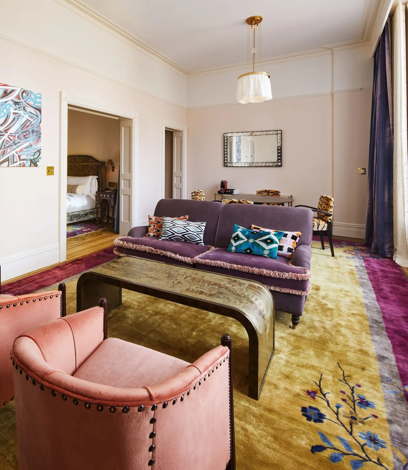 Chelsea Grand Loft at Hotel Chelsea: a living room with a purple sofa, pink armchairs and a long brass-topped coffee table, with a bedroom visible through an open double doorway
