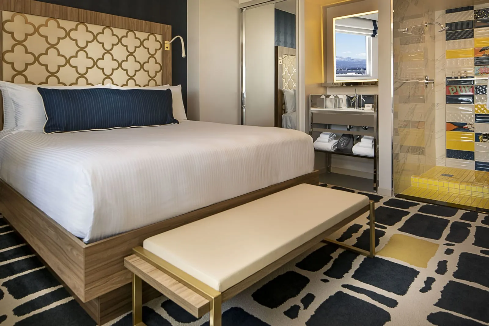 Blanca King at SAHARA Las Vegas: a king bed with a quatrefoil headboard and a navy cushion, an upholstered bench at its foot, and an open vanity beside a shower tiled in blue, yellow and white pop-art panels