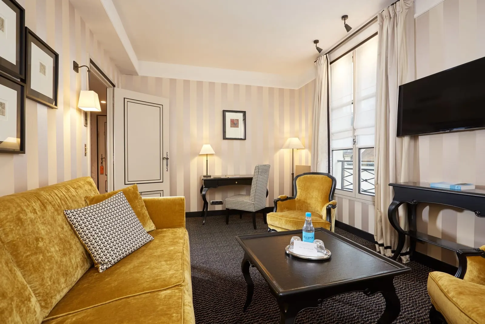 Junior Suite at Le Pavillon de la Reine: the sitting room, with a yellow velvet sofa and armchairs, a coffee table, a writing desk and a television on the wall