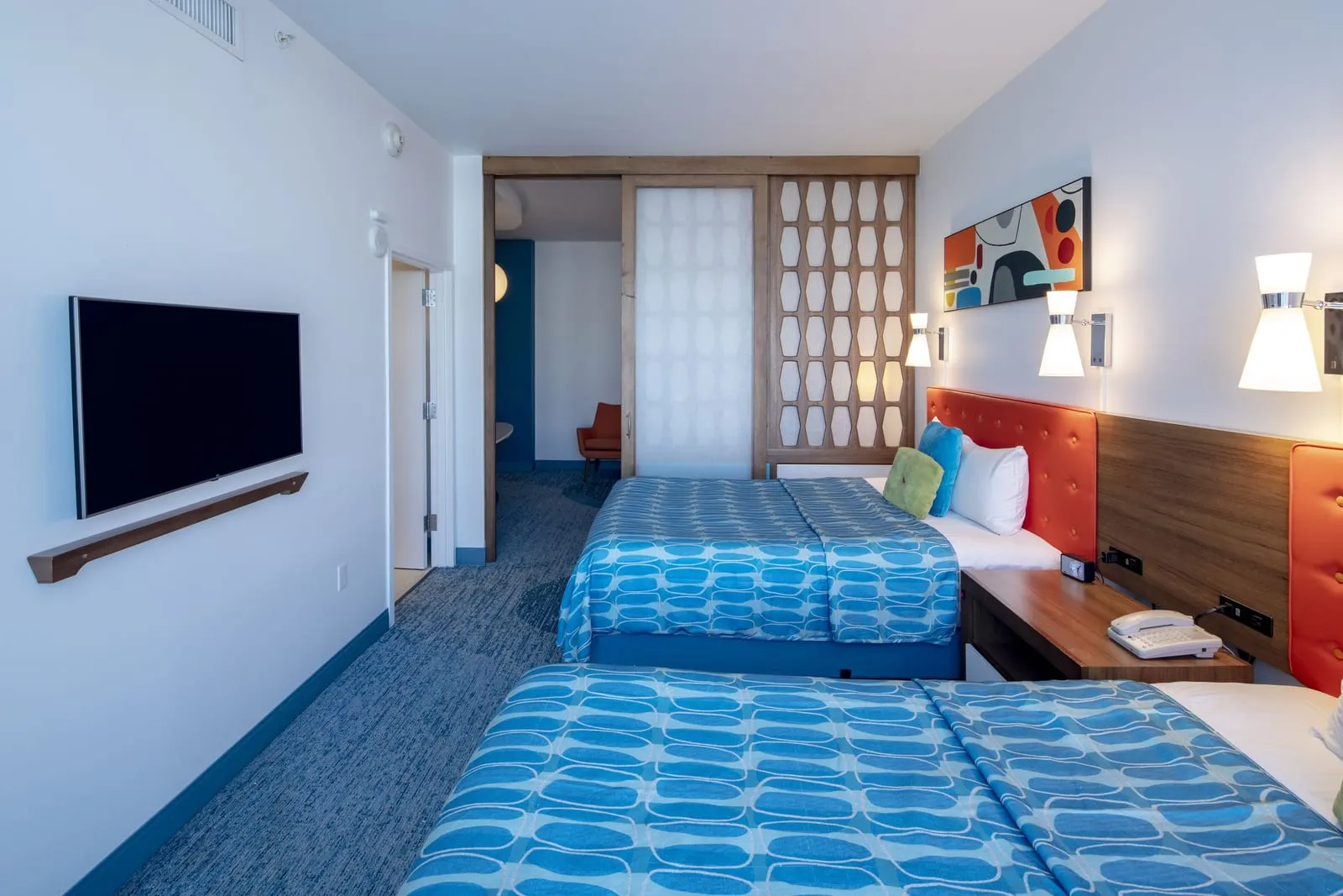 Poolside Family Suite Exterior Entry at Universal Cabana Bay Beach Resort: two queen beds with orange headboards, a wall-mounted television and the sliding partition half open onto the living room and an orange armchair