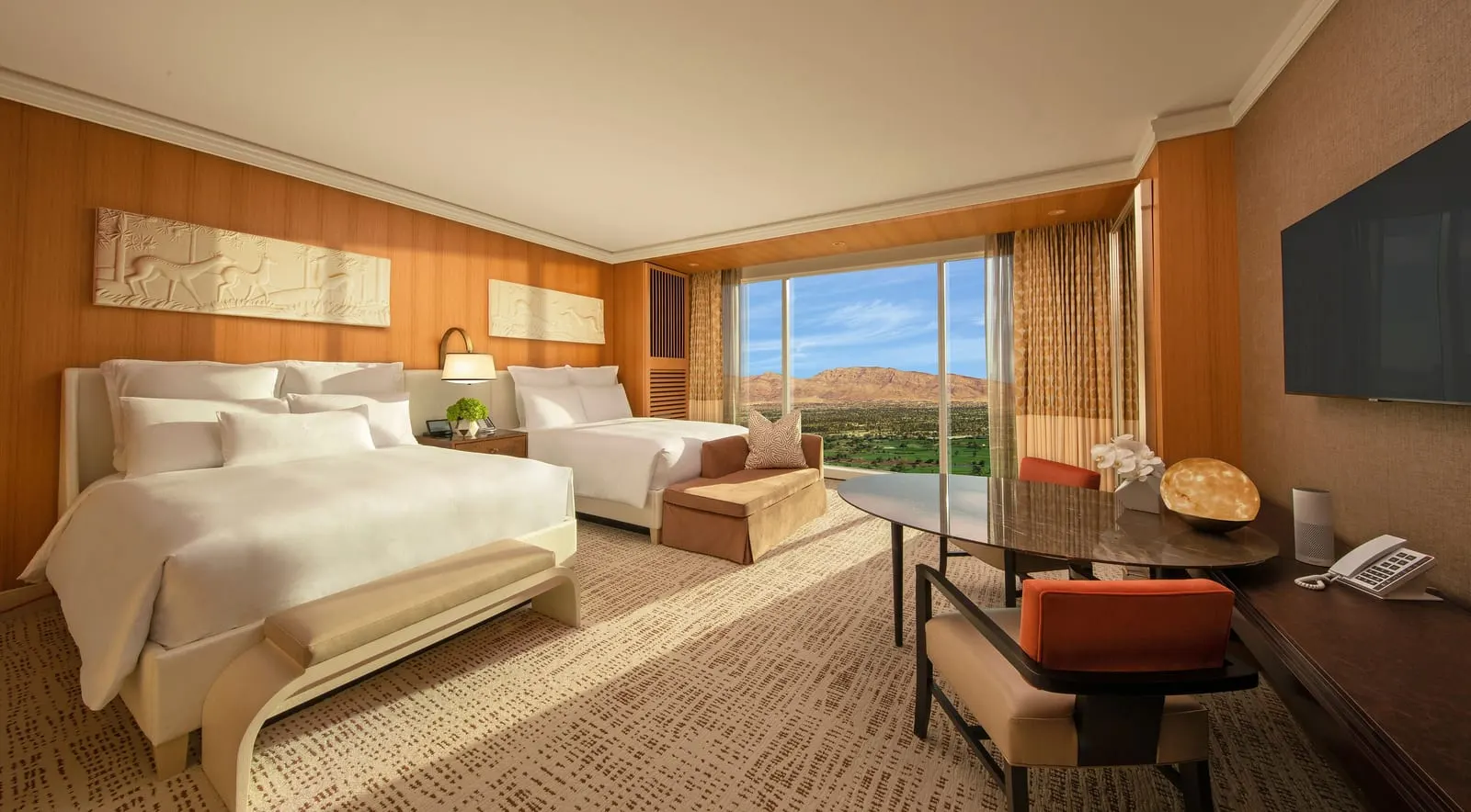 Wynn Resort Two Queens at Wynn Las Vegas: two queen beds below carved relief panels, a bench at the foot of one, a small sofa and an oval table with two orange-backed chairs by a floor-to-ceiling window