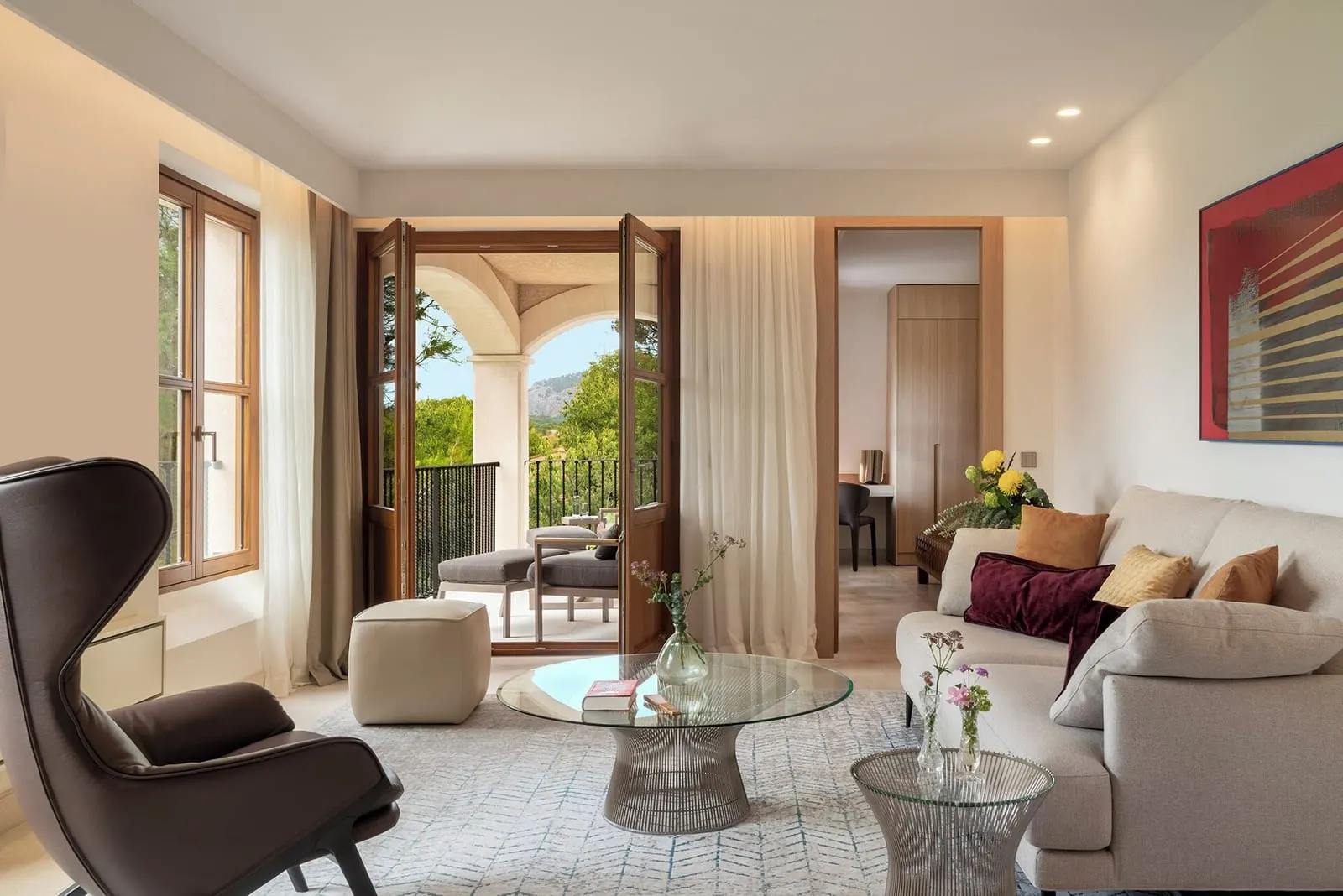 Garden Suite im Castell Son Claret: ein Wohnzimmer mit einem cremefarbenen Sofa, einem Ledersessel und einem gläsernen Couchtisch und Fenstertüren mit Rundbogen zu einem Balkon mit Liegen und Bäumen dahinter